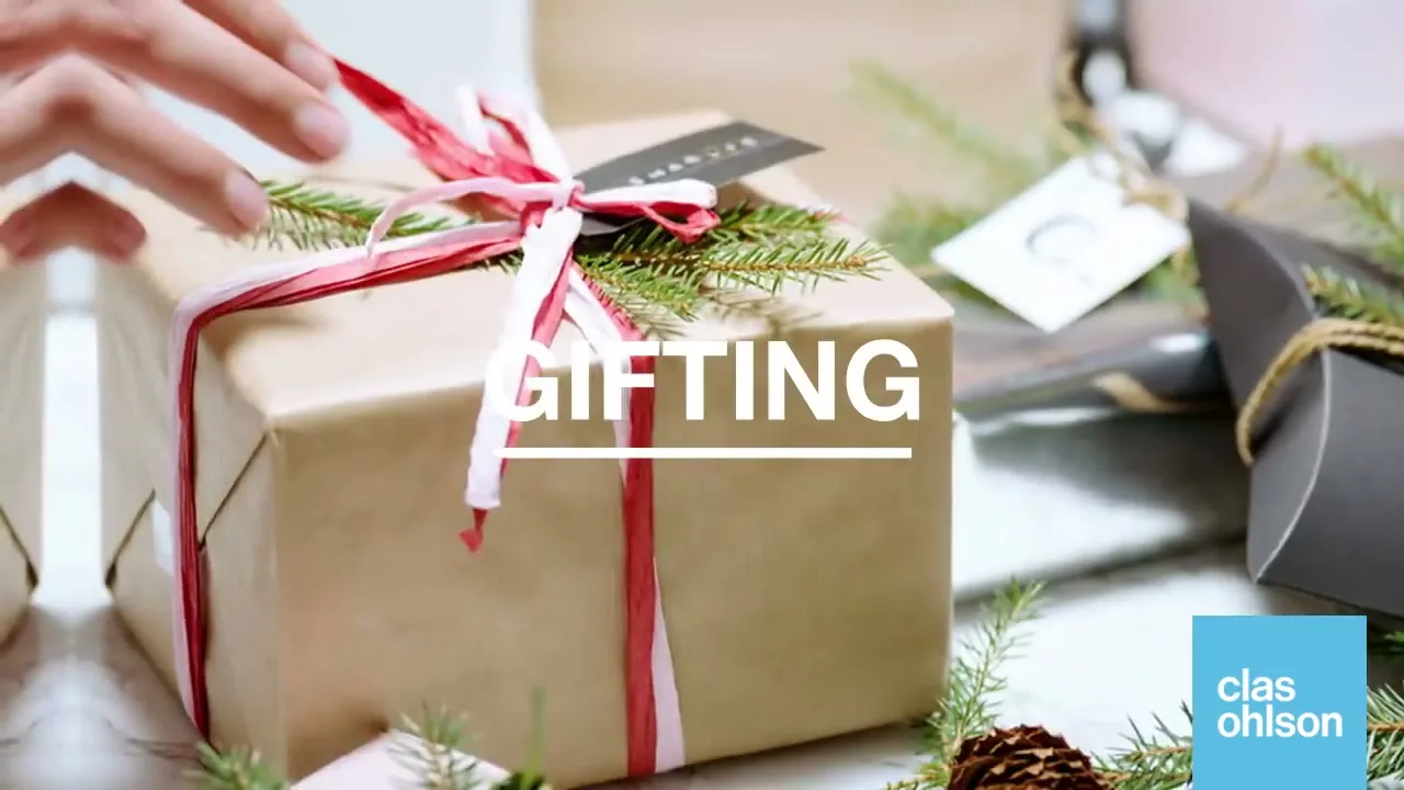 A person's hands are seen tying a red and white ribbon around a brown paper gift box, surrounded by other gifts and pine needles on a table.