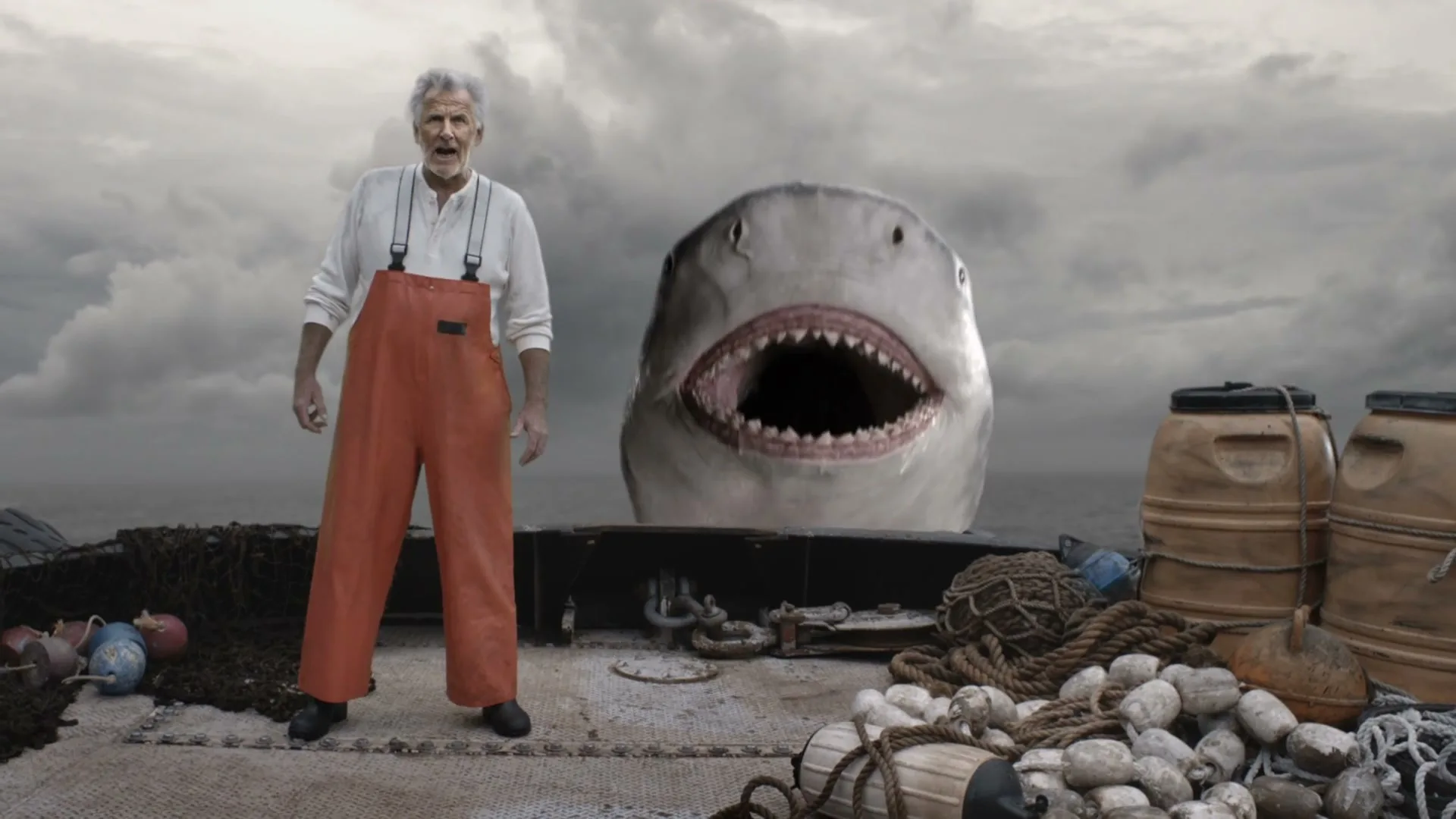 An old fisherman in orange waders stands on a boat deck, looking at the camera, as a large shark with an open mouth emerges from the water behind him under a cloudy sky.