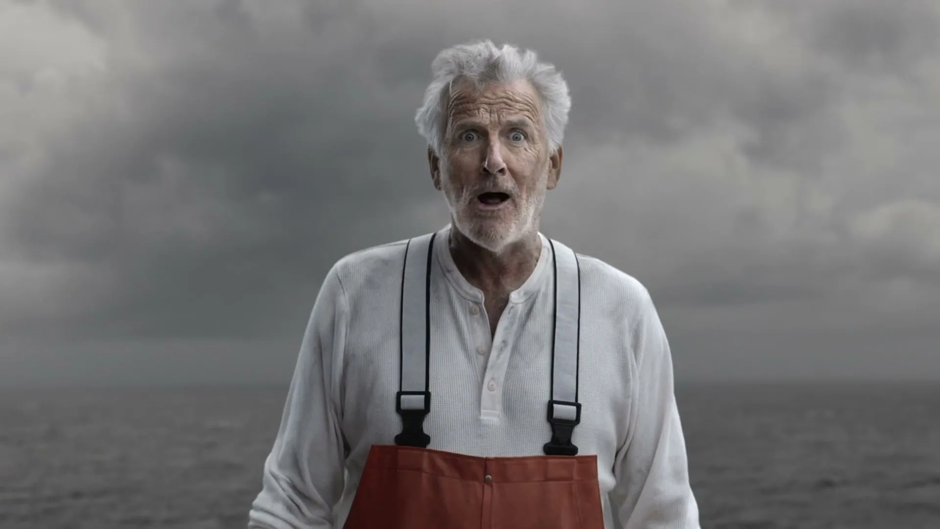 A grizzled old fisherman with white hair and a beard, wearing a white shirt and orange overalls, looks directly at the camera with a surprised expression, standing in front of a stormy sea and sky.