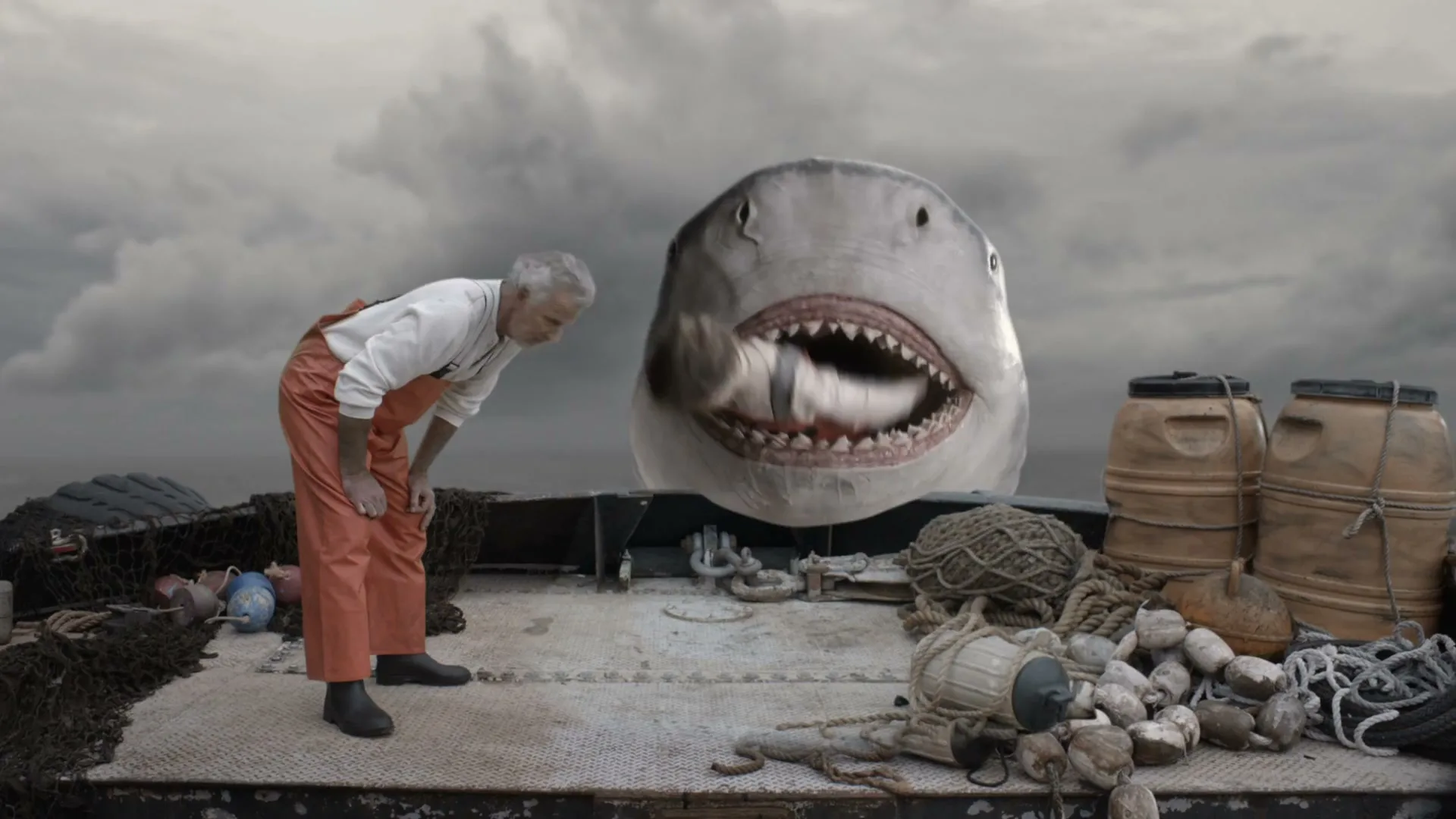 An old fisherman in orange waders bends over on a boat, looking at a giant shark with its mouth open, on a cloudy day.