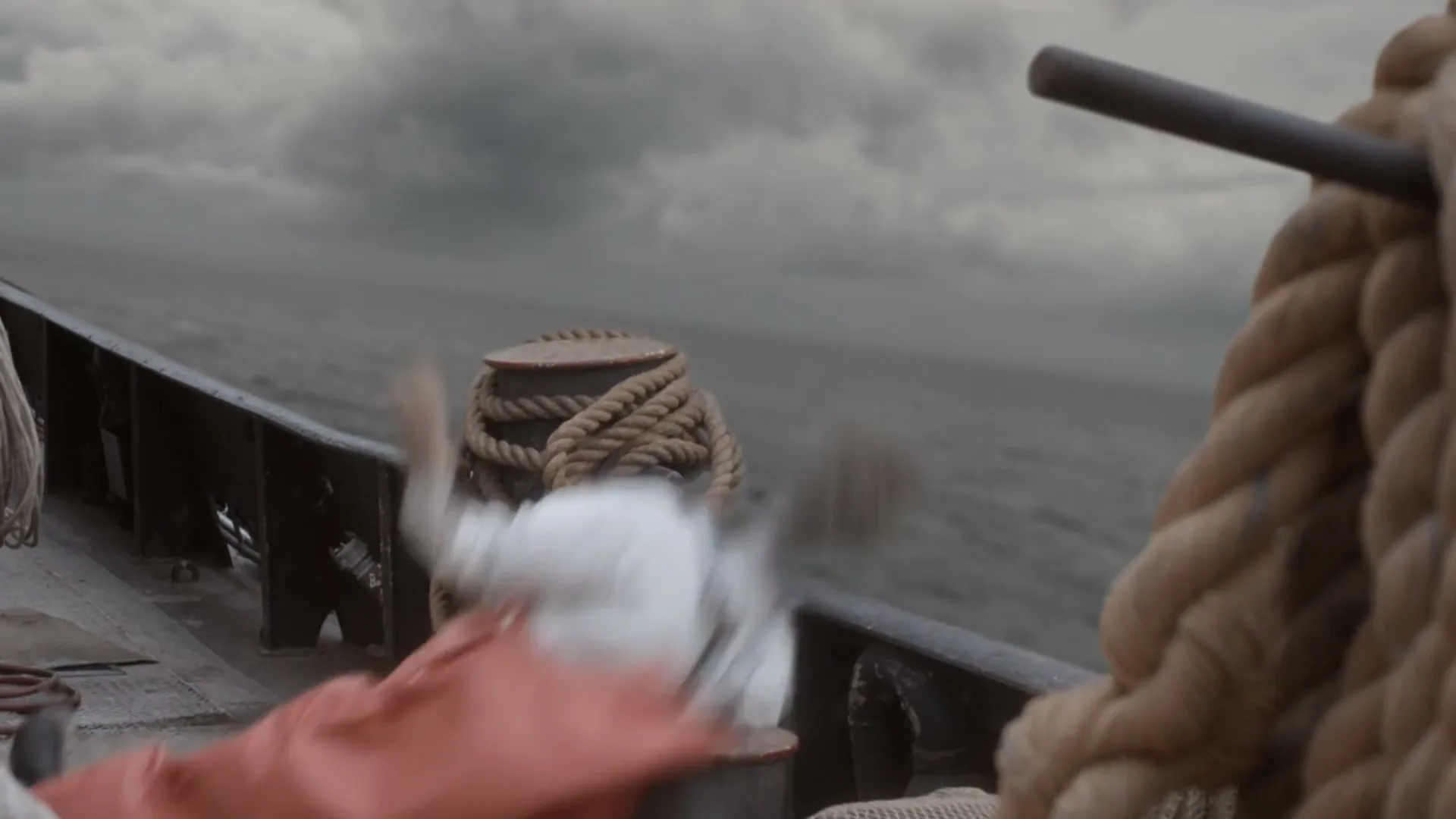 A fisherman in a white shirt and orange gloves is seen in a blur as he falls on the deck of a fishing boat, with ropes and a stormy sea in the background.