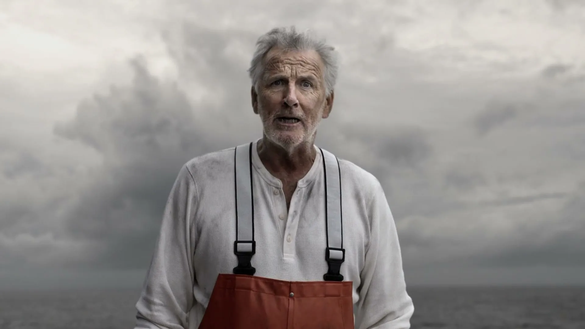 A grizzled old fisherman with a white beard and red overalls looks directly at the camera with a surprised expression, against a stormy sea and sky.