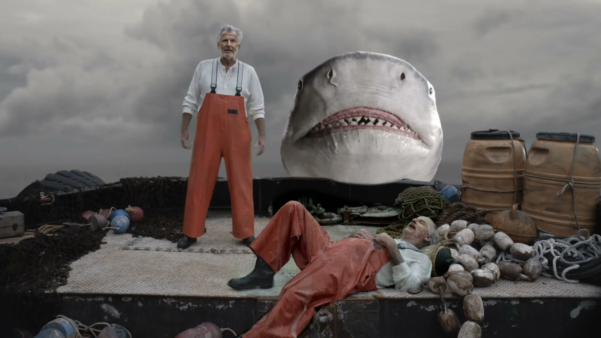 Two fishermen on a boat, one standing and one lying down, with a giant shark head behind them under a cloudy sky.