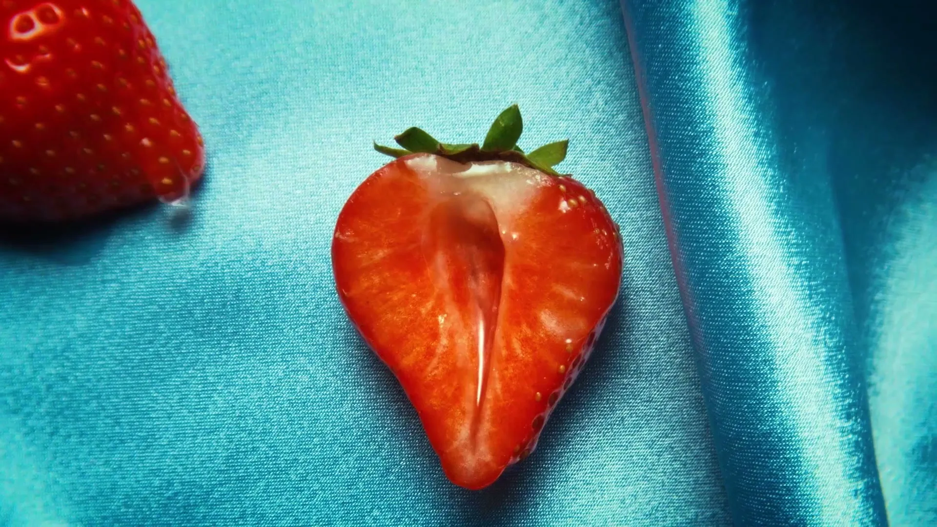A close-up shot of a halved strawberry with a white liquid inside, resting on a blue satin fabric.