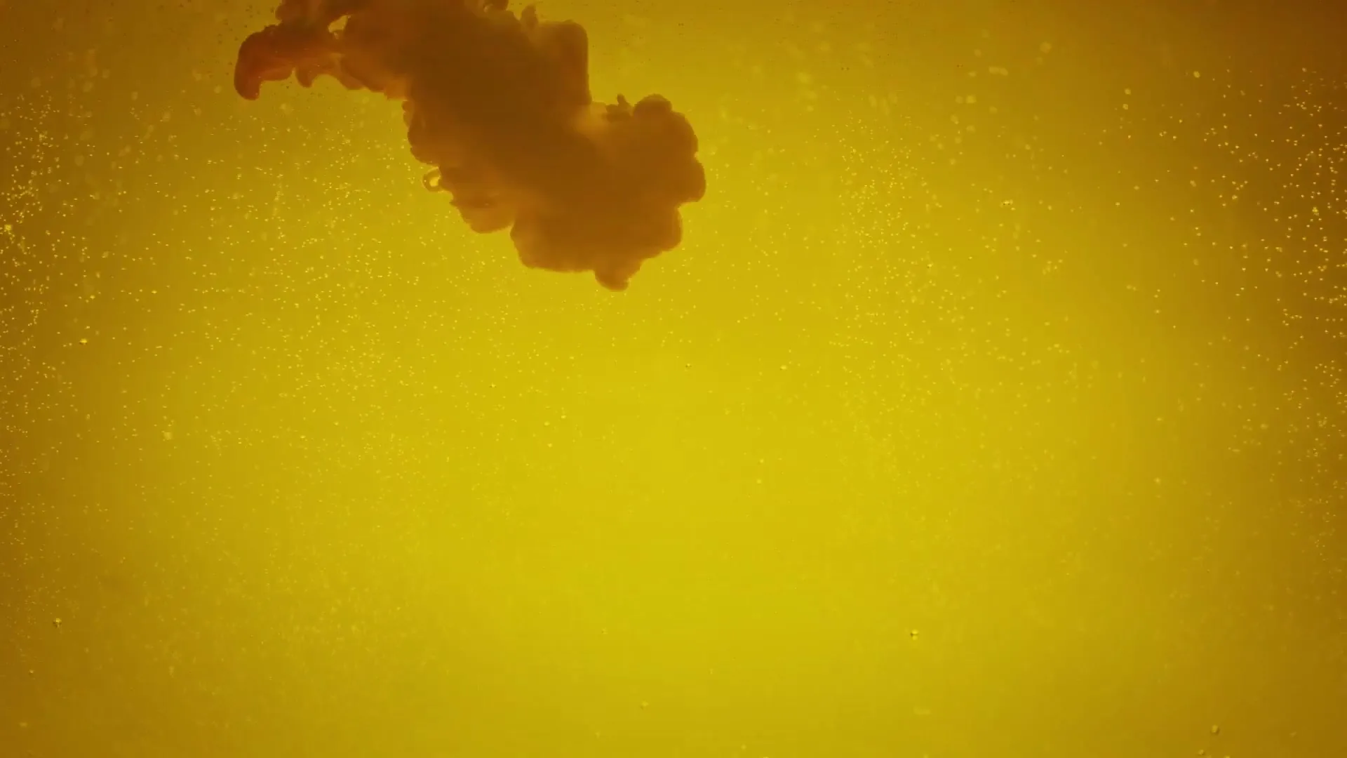 A thick, orange liquid slowly disperses into a yellow liquid, creating a cloudy, swirling effect with tiny bubbles.