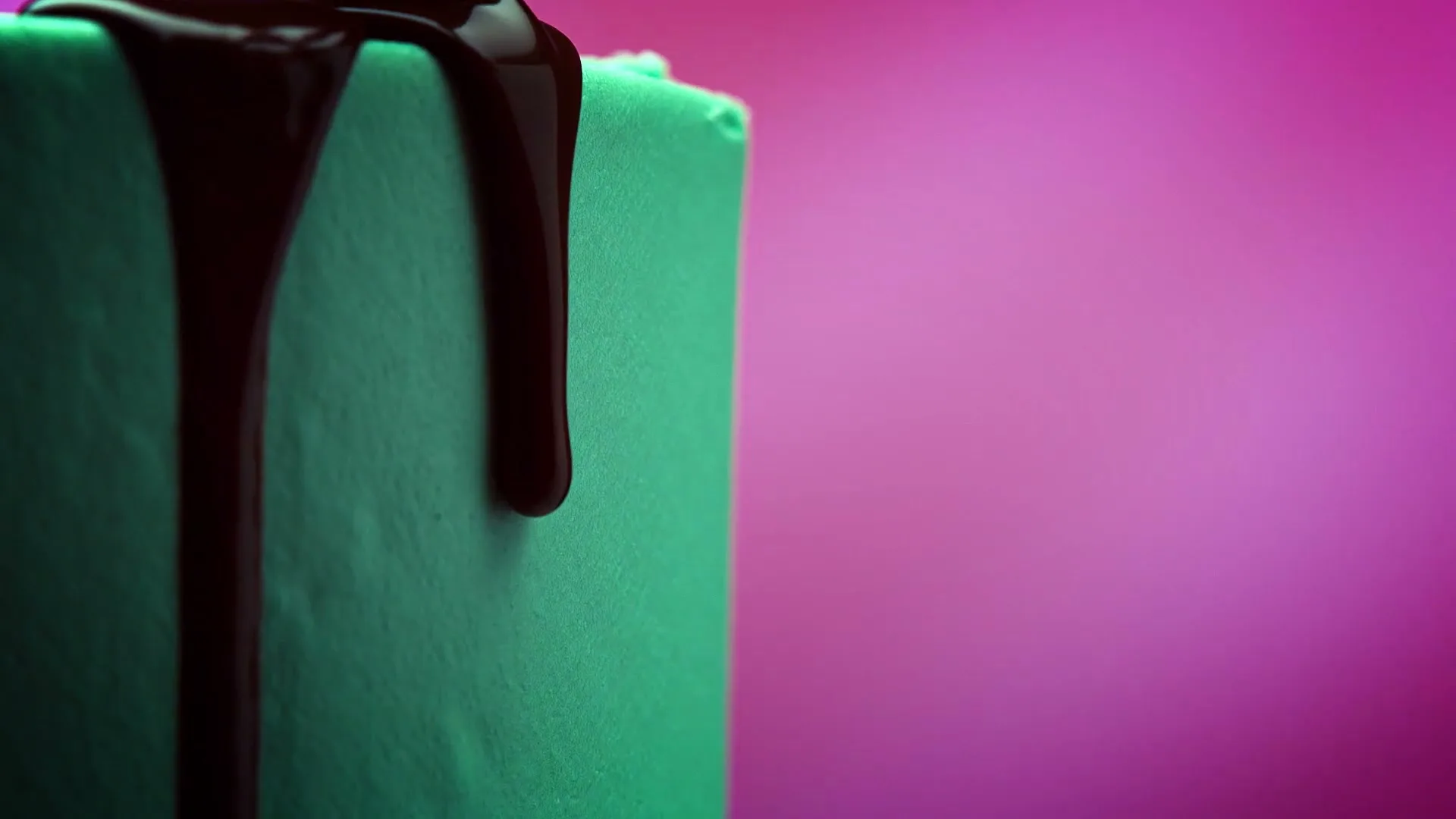 Chocolate sauce drips down the side of a green cake against a vibrant pink background, creating a visually appealing dessert scene.