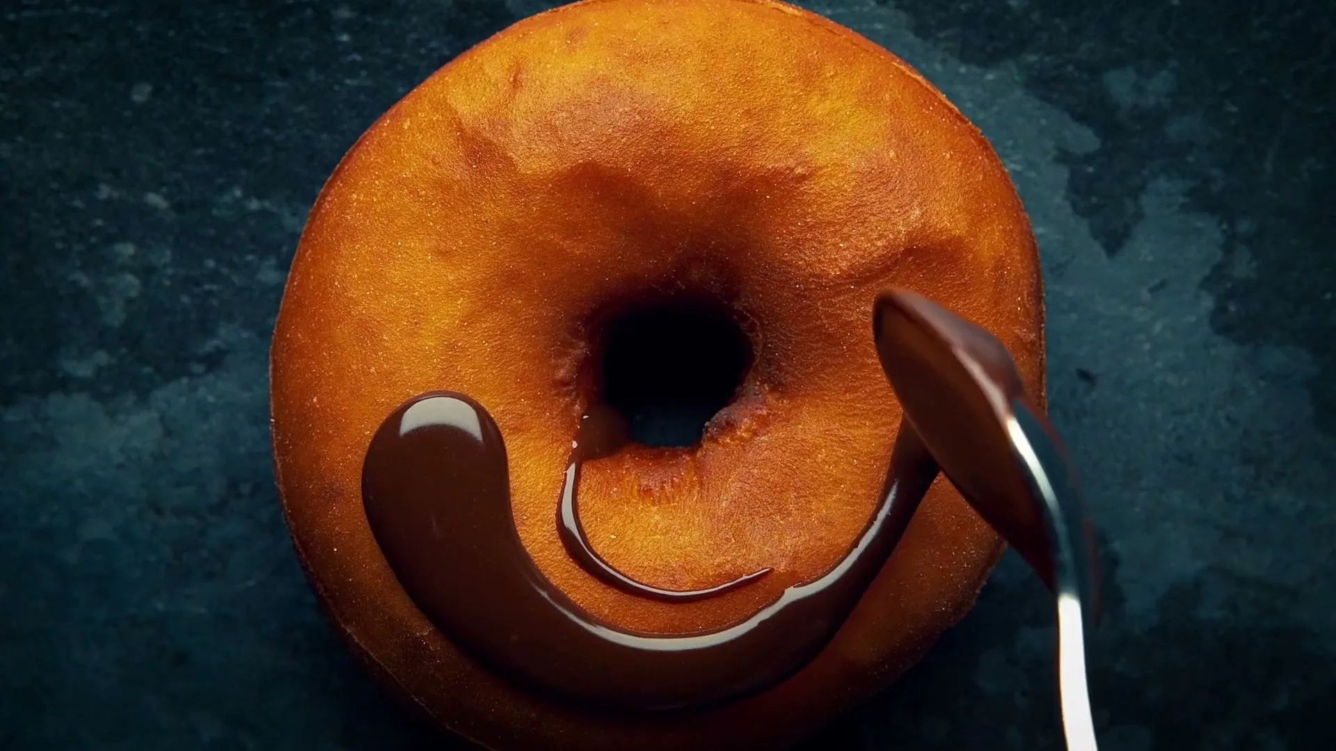 A spoon drizzles thick chocolate sauce onto a golden-brown donut, creating a swirling pattern on a dark, textured surface.