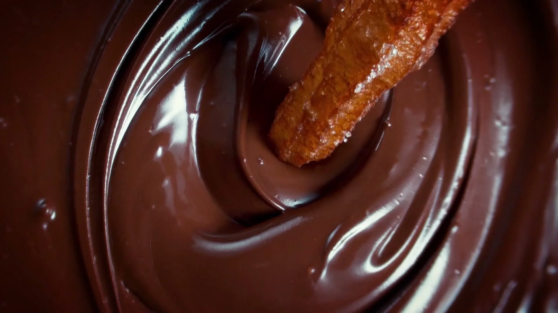 A churro is dipped into a swirling pool of rich, melted chocolate, creating ripples and reflections on the surface.