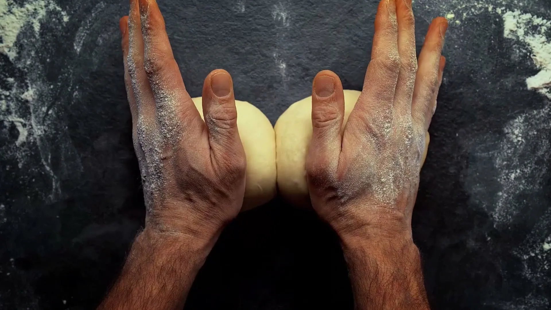 Two male hands, covered in flour, knead a piece of dough on a dark, flour-dusted surface, pressing it together.