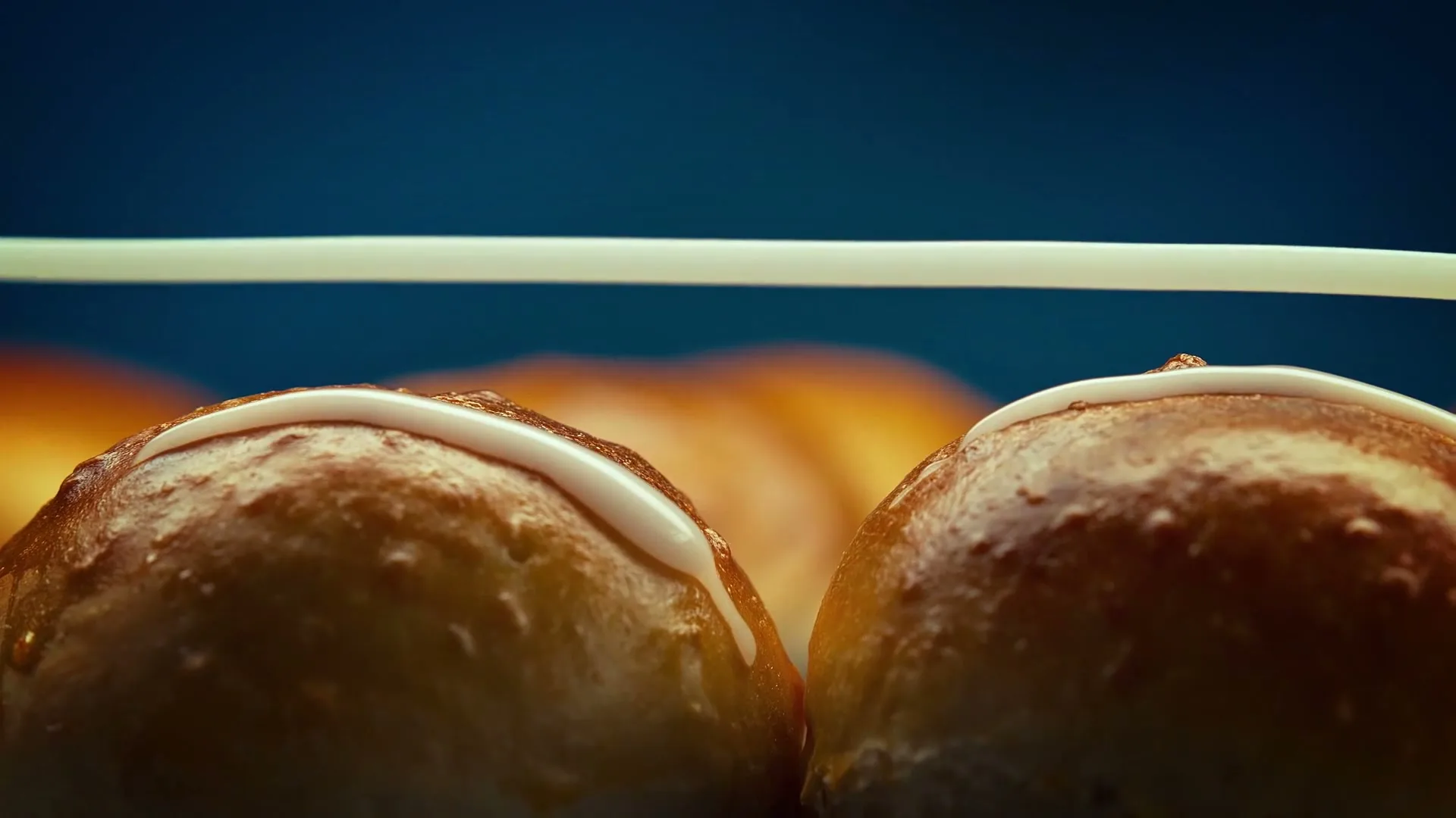 A close-up shot shows two buns with white icing being drizzled over them, against a blurred blue background.