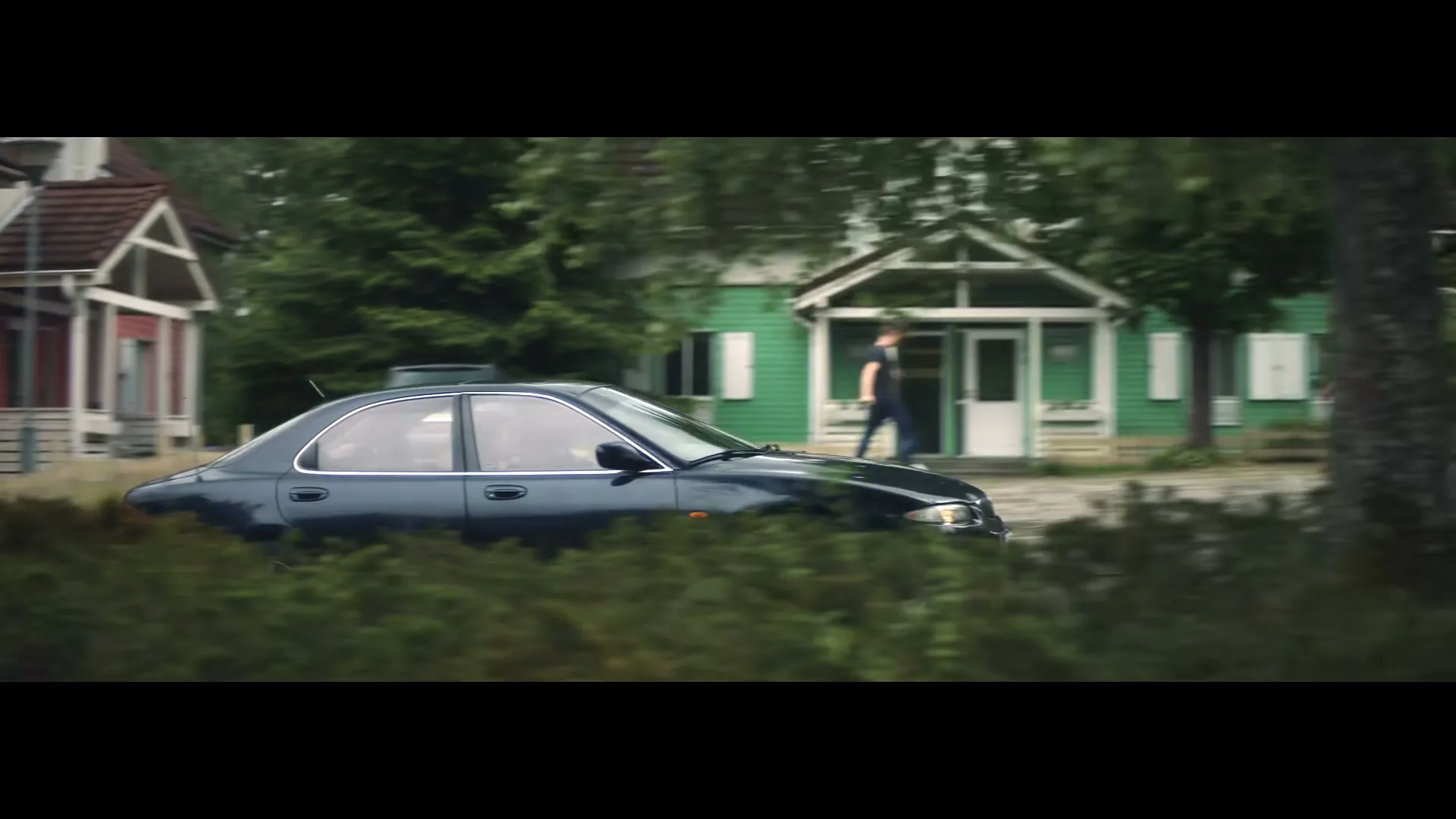 A dark blue sedan drives past a green house with a man walking out, seen through bushes in a suburban neighborhood.