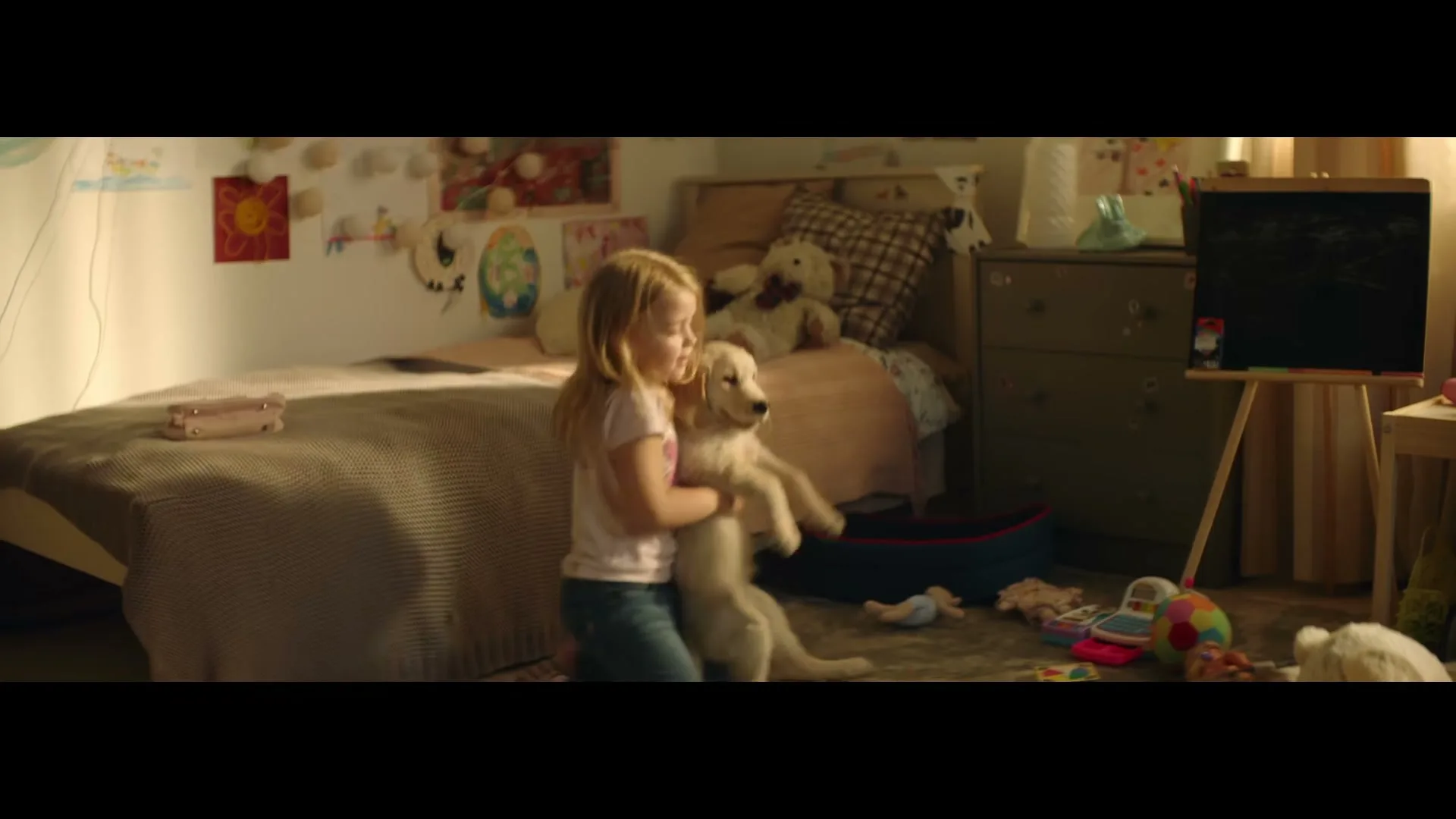 A young blonde girl in a pink shirt and jeans holds a golden retriever puppy in a sunlit bedroom, then kneels down with it.