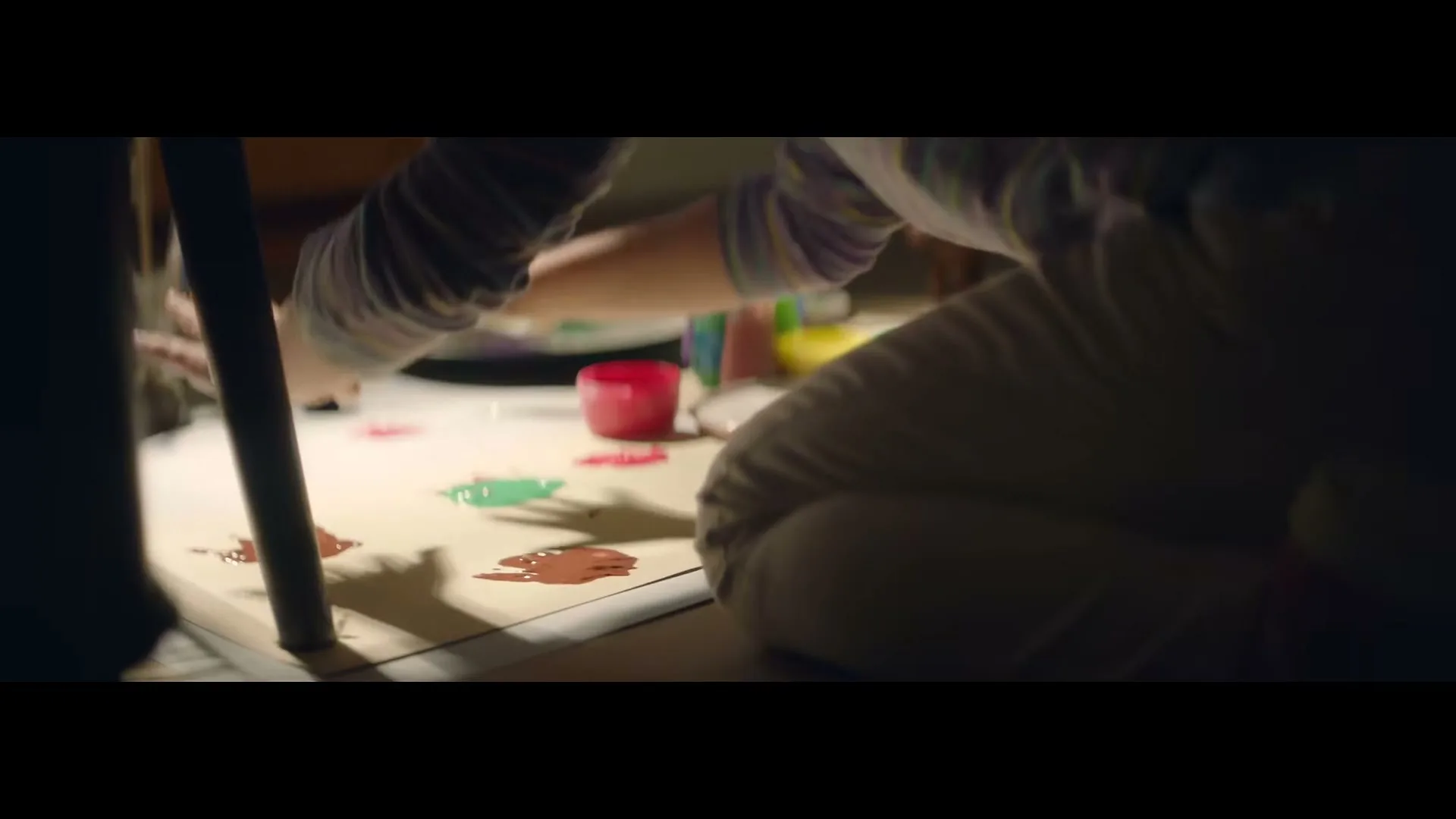 A child in a striped shirt and grey pants kneels on the floor, finger painting with various colors on a white sheet of paper in a dimly lit room.