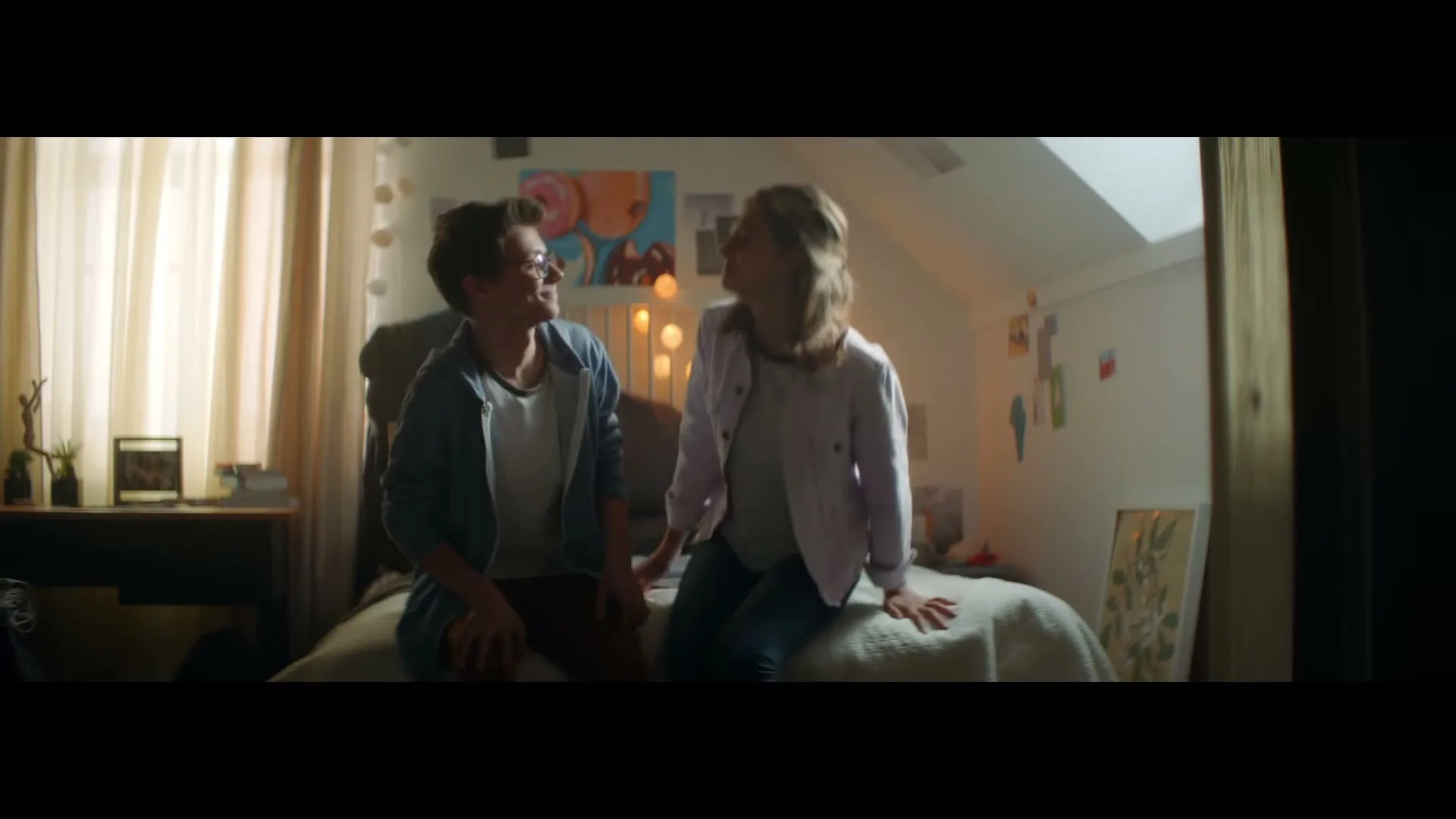A young male and female sit on a bed in a cozy bedroom, looking at each other and smiling warmly.
