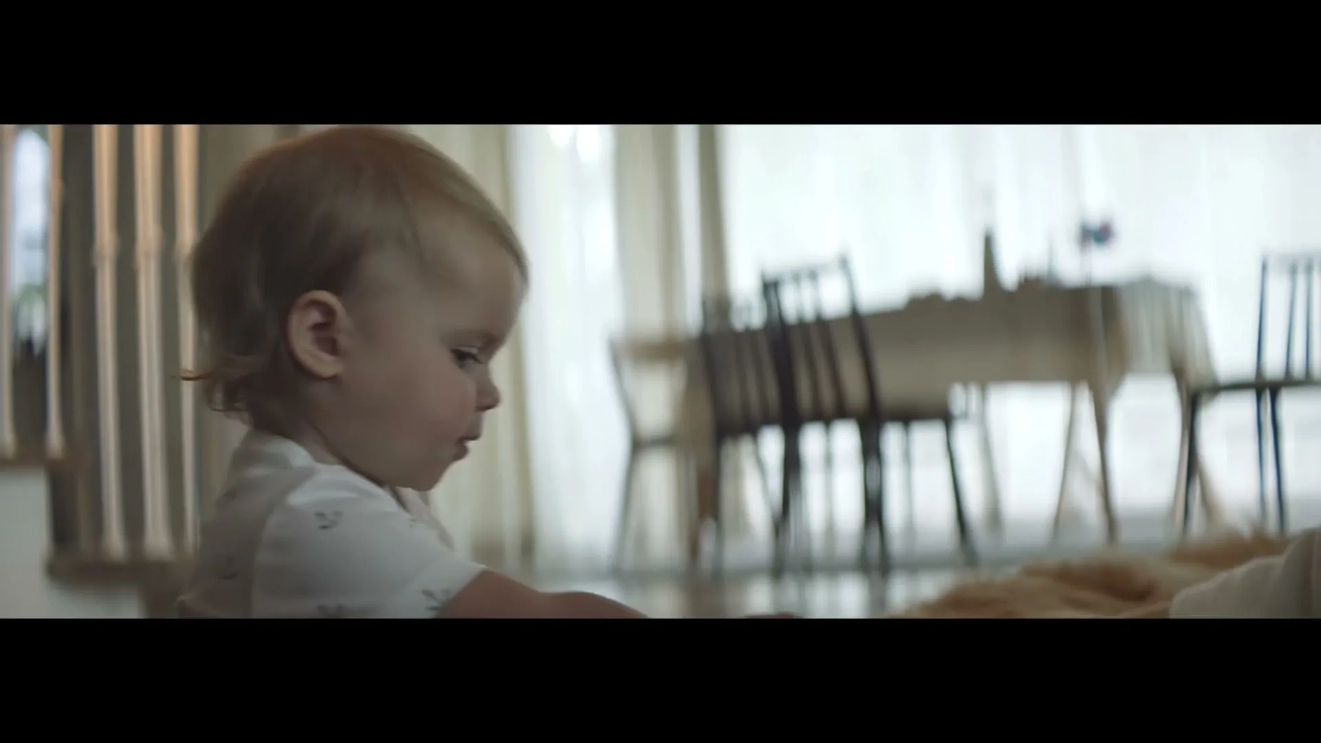 A baby with blonde hair in a white onesie sits on a wooden floor, looking down at a blue toy in their hands in a bright room.