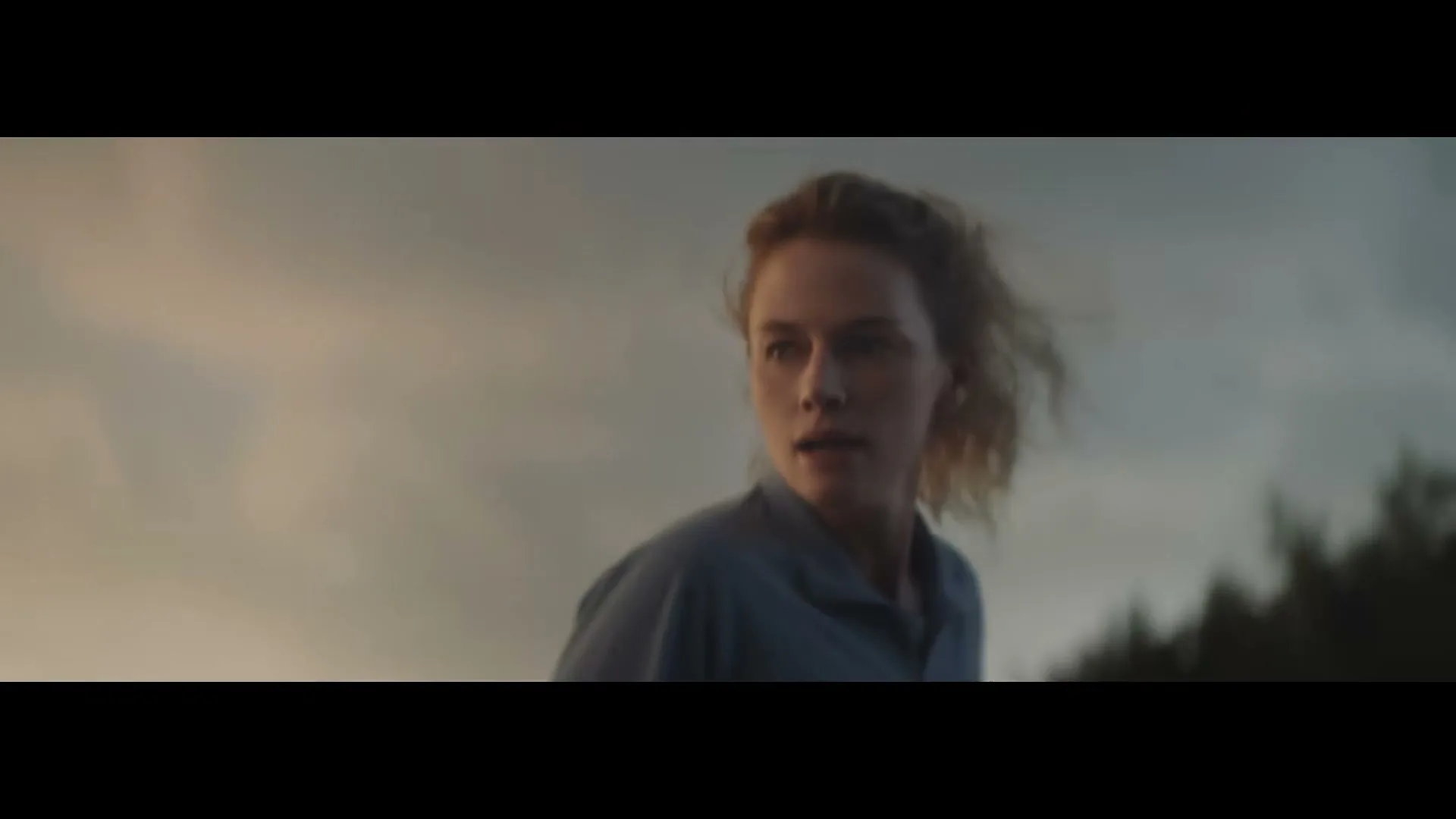 A young woman with blonde hair and a blue shirt looks over her shoulder with a concerned expression, as the wind blows her hair, against a cloudy sky.