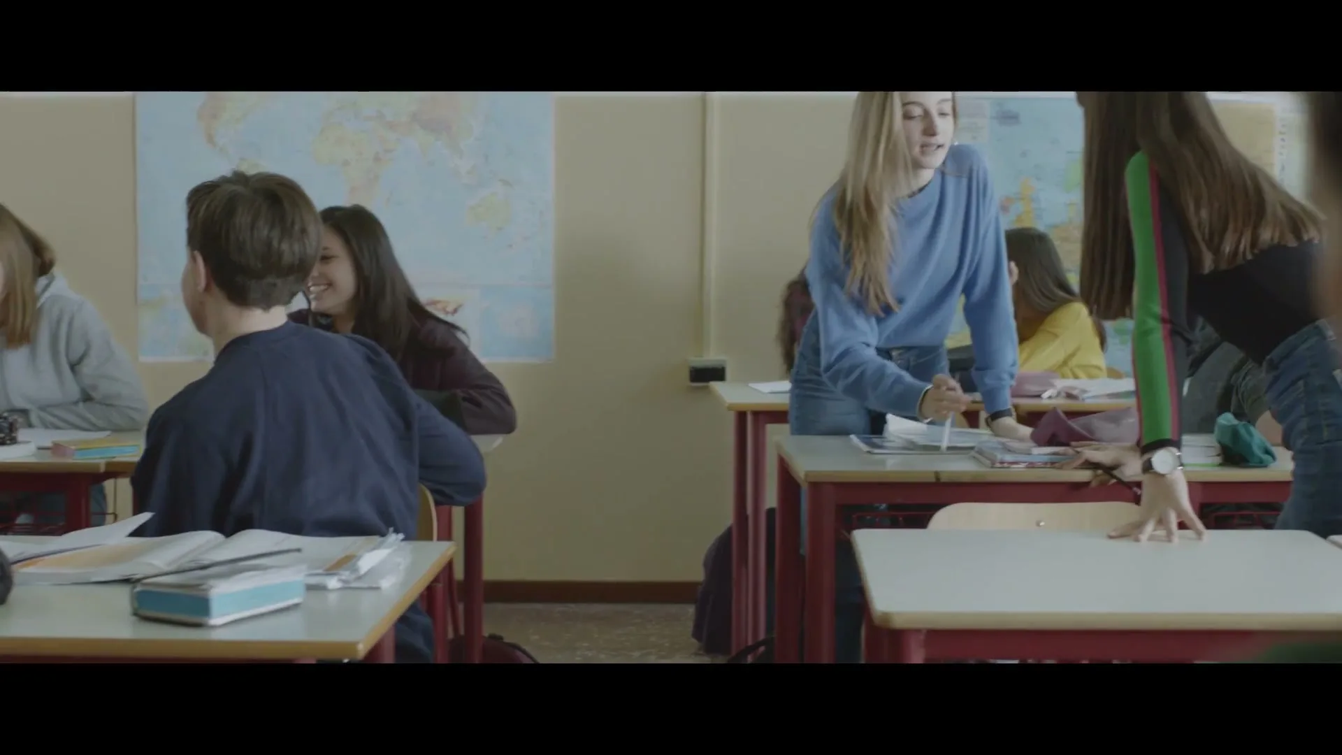 A classroom of high school students are talking and laughing, with two girls standing by a desk in the foreground, and a boy in the midground.