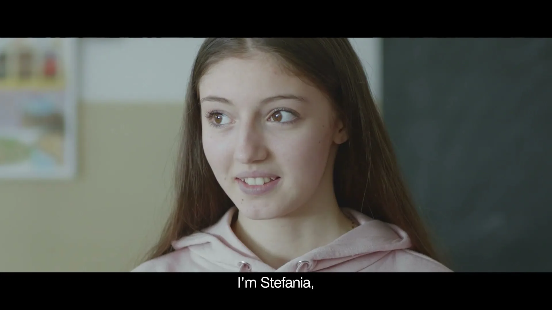 A young female student with long brown hair and a pink hoodie speaks to someone off-camera in a classroom.