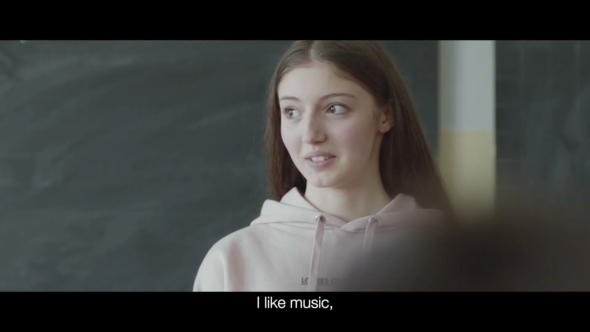 A young female student with long brown hair, wearing a pink hoodie, smiles and speaks in a classroom with a blackboard behind her.