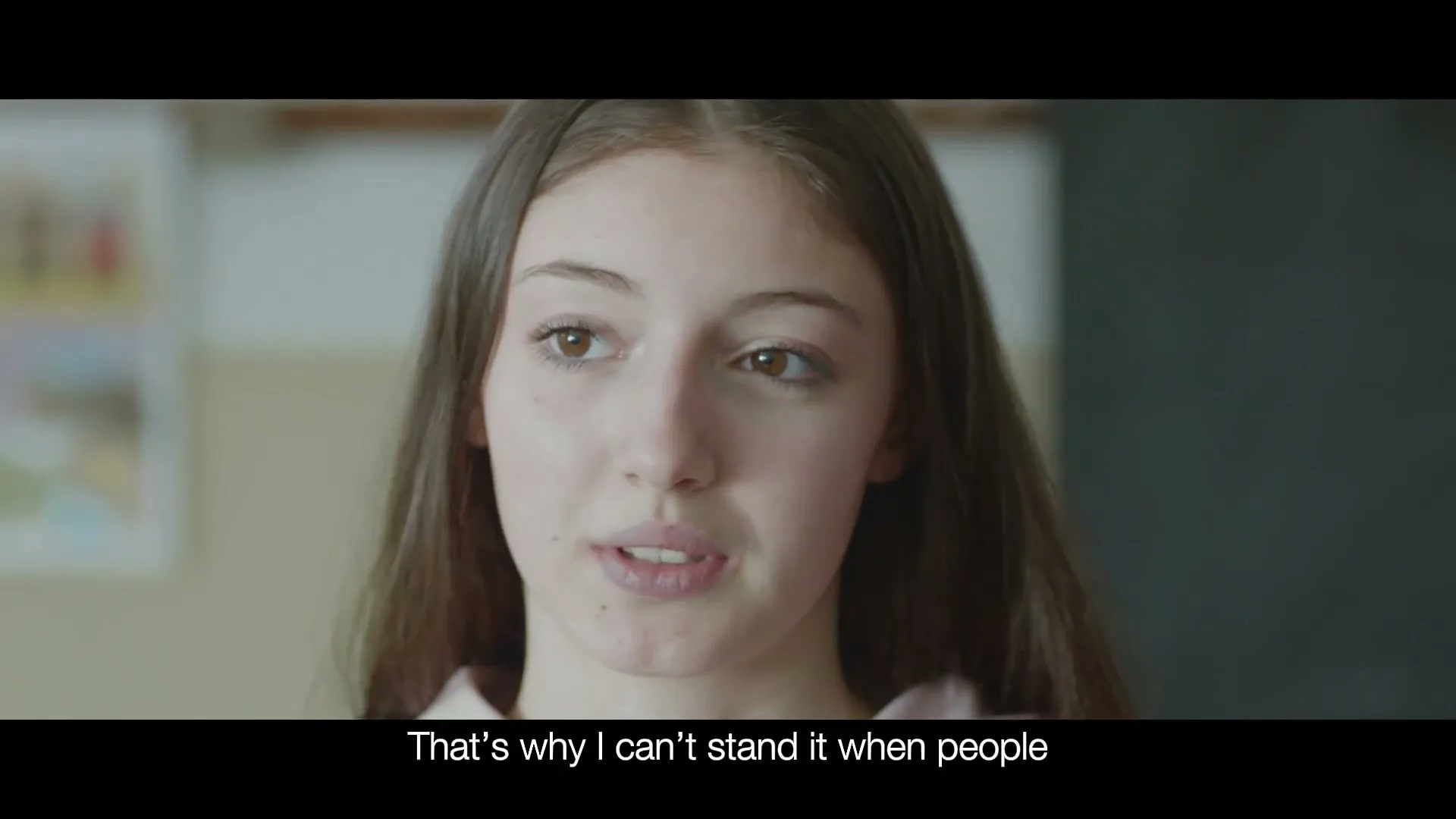 A young female with long brown hair and a light pink top speaks directly to the camera in a classroom setting, expressing discomfort.