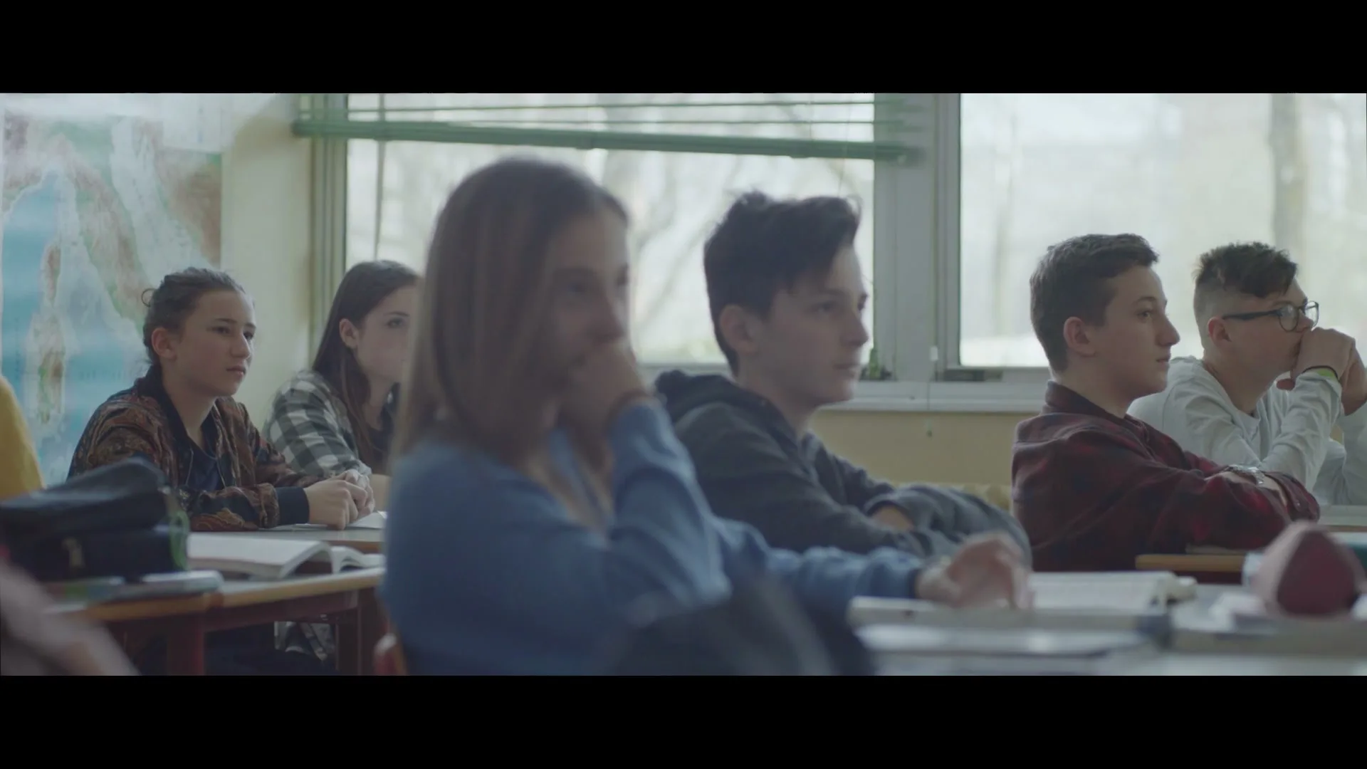 A group of diverse teenagers sit at desks in a classroom, listening intently to someone off-screen, with natural light streaming through windows.