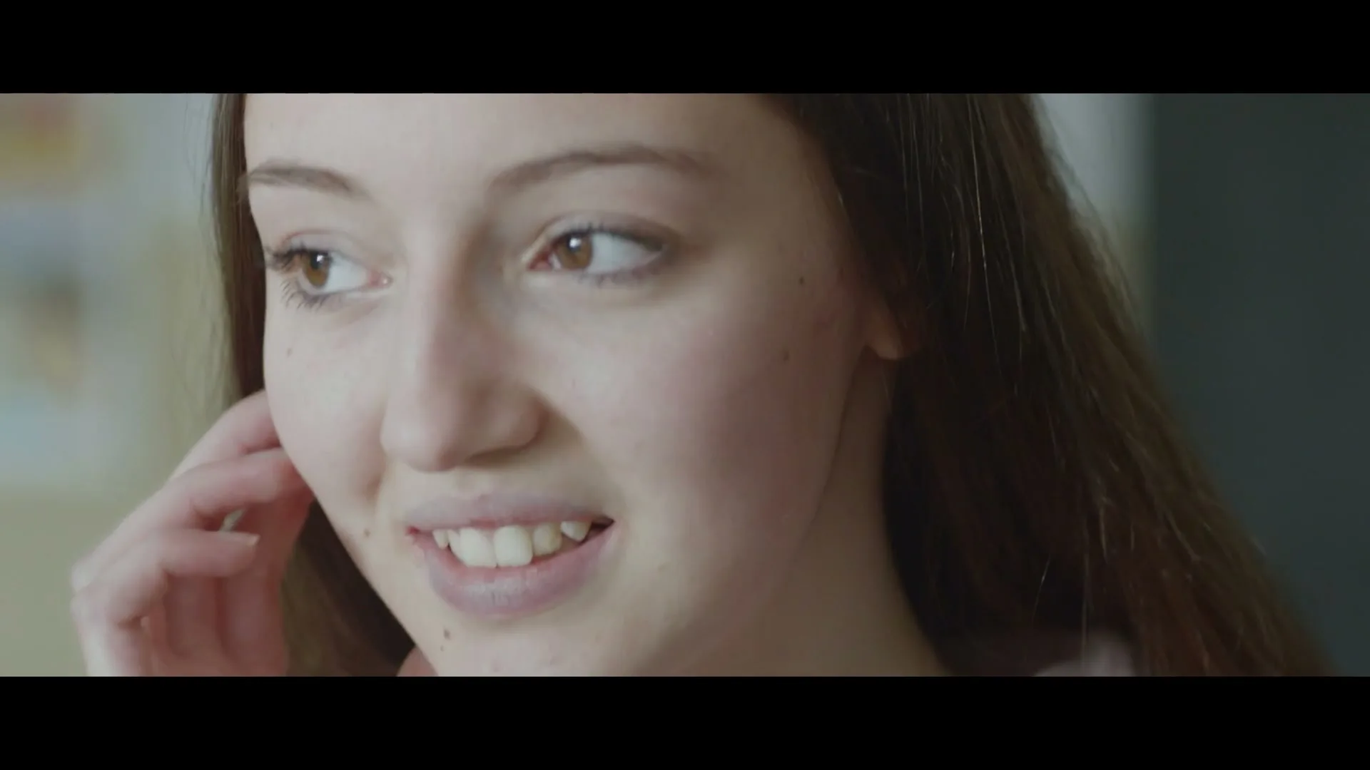 A young female with long brown hair and a light pink top looks down and then up, speaking softly in an interior setting.