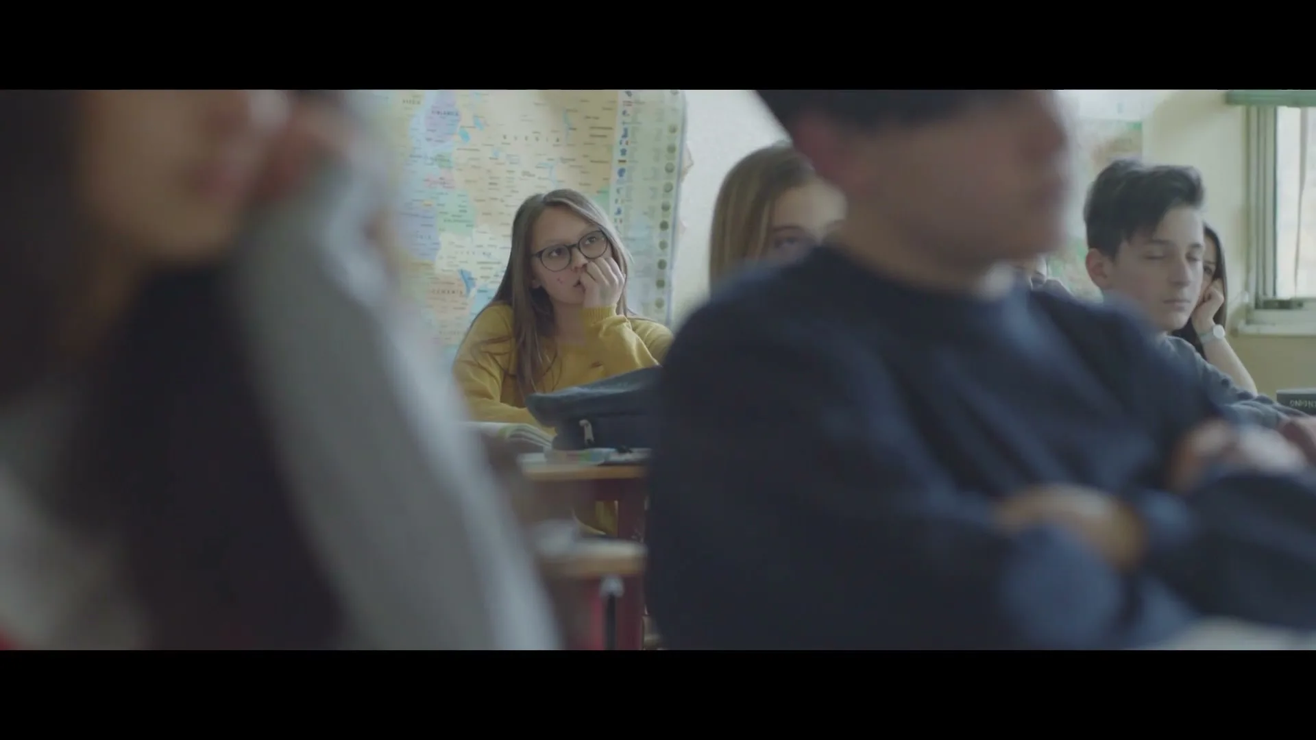 A young girl with glasses and a yellow top looks bored in a classroom, surrounded by other students, with a map on the wall.