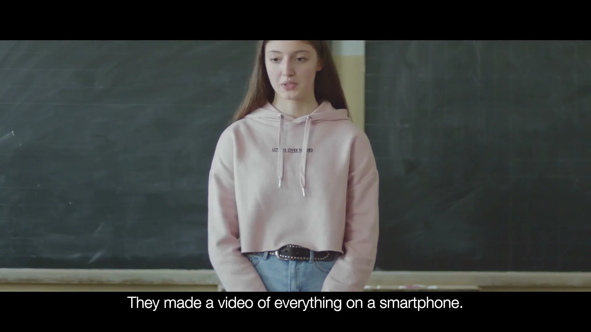 A young female student with long brown hair, wearing a pink hoodie and jeans, stands in front of a blackboard in a classroom.