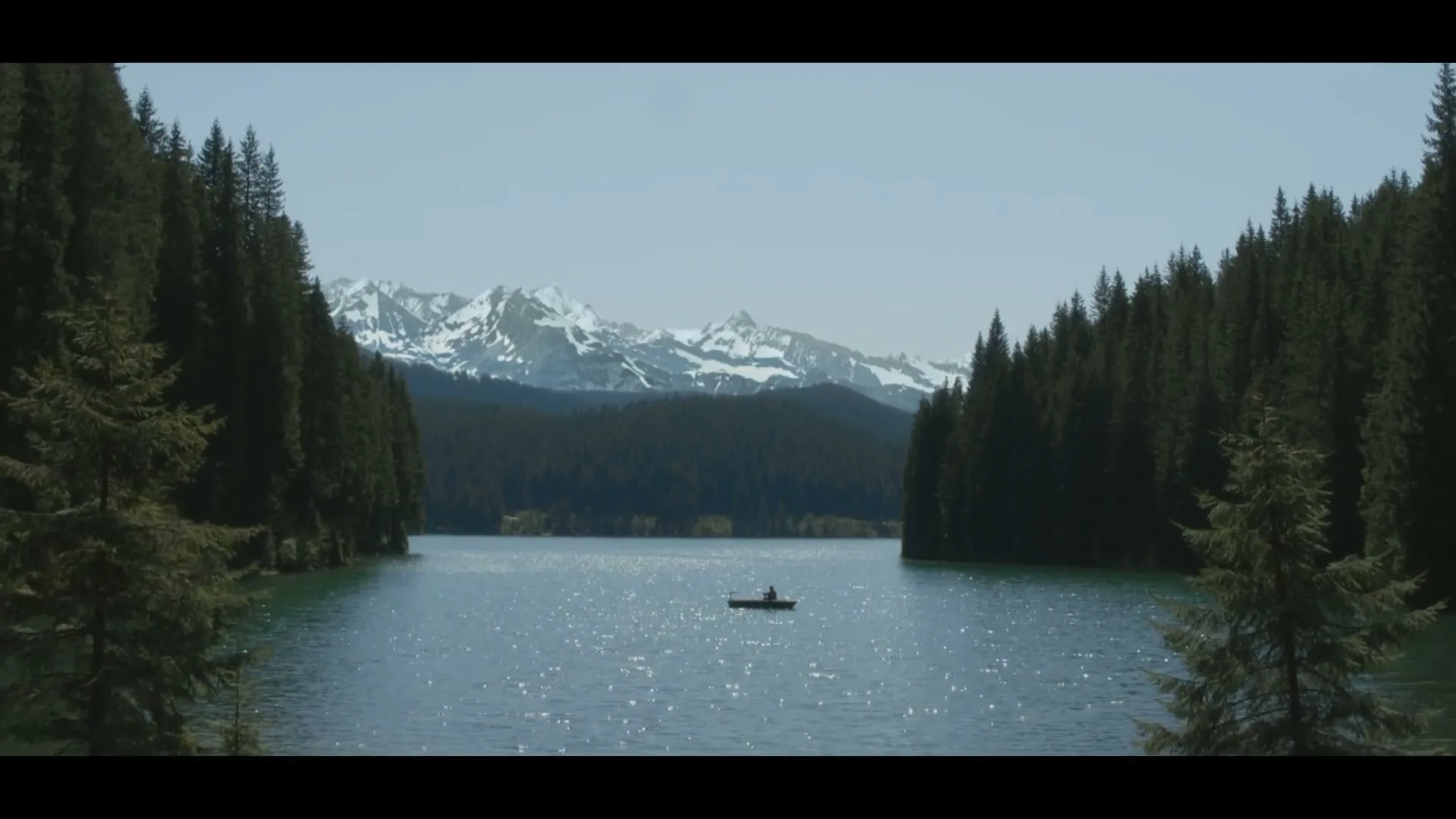 A lone person in a small boat fishes on a sparkling lake surrounded by dense pine forests and snow-capped mountains under a clear sky.