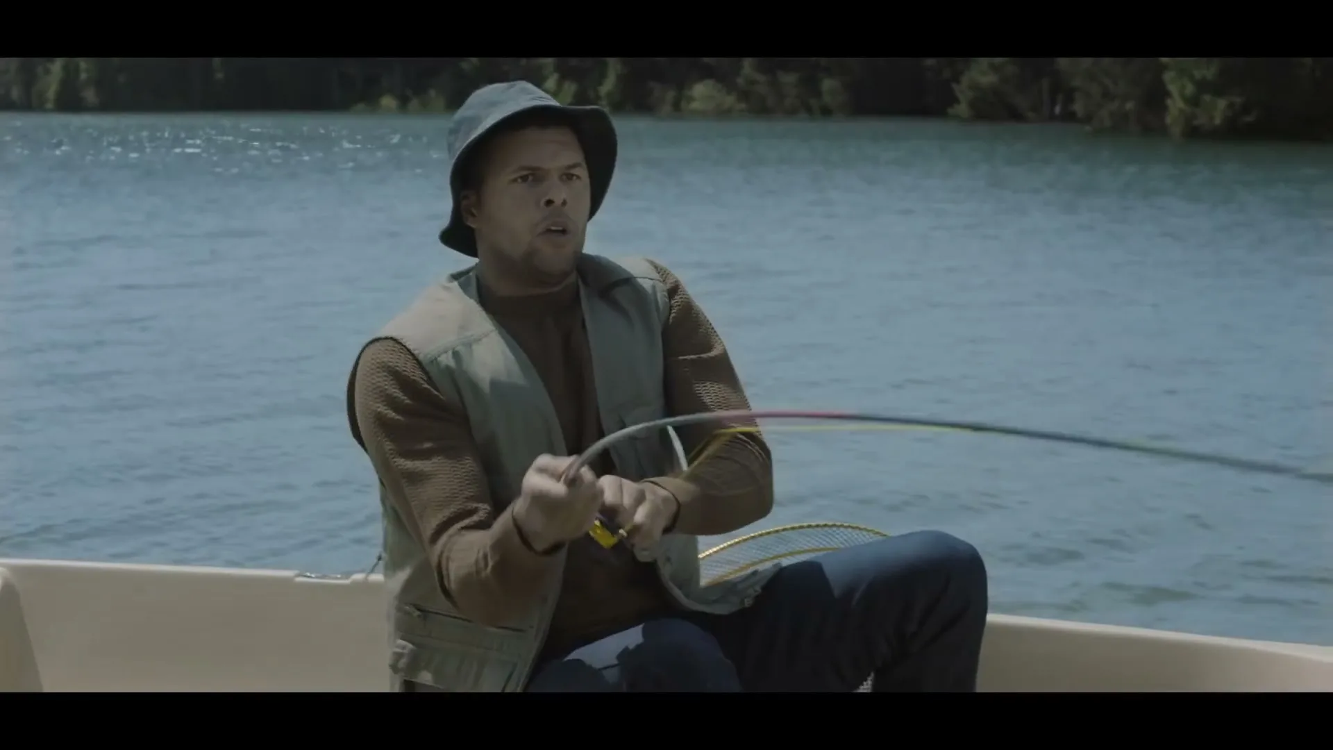 A man in a bucket hat and fishing vest struggles to reel in a fish from a small boat on a lake during the day.