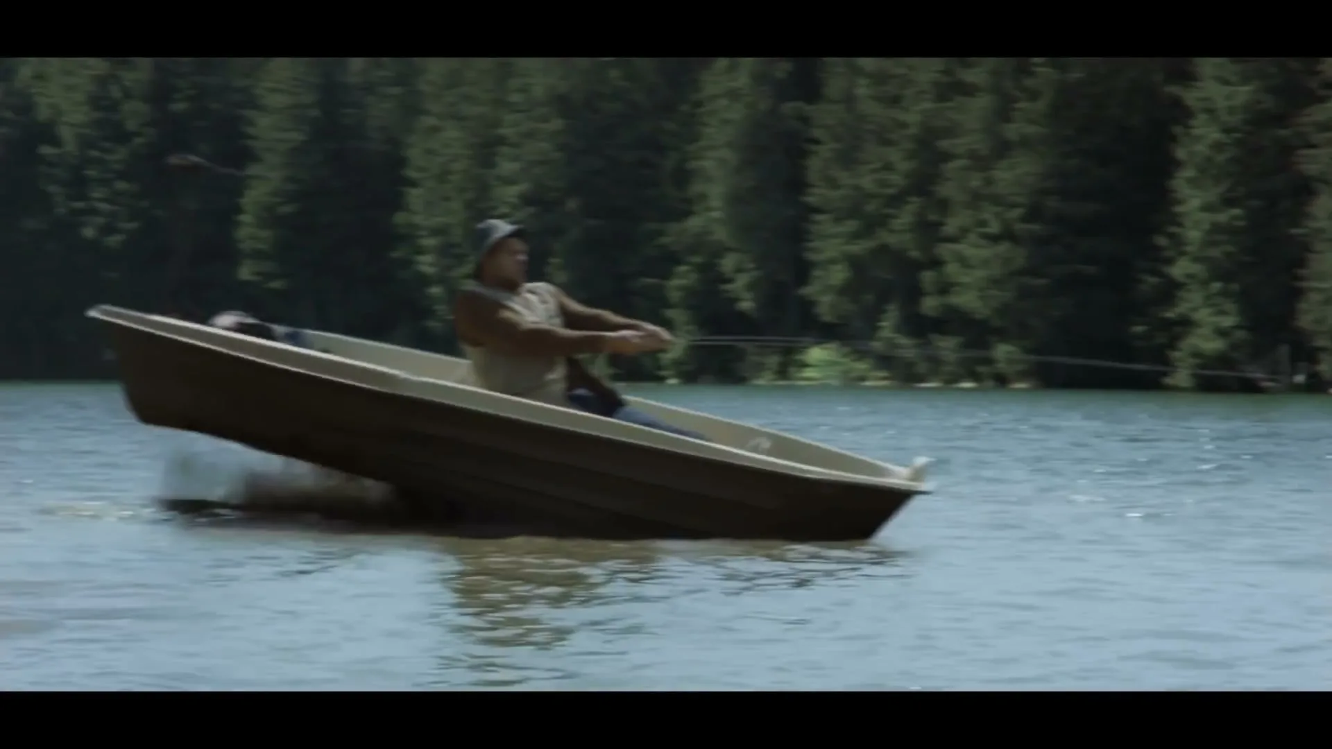 A man in a fishing boat on a lake is pulled backward by a strong fish, causing the boat to tilt precariously.