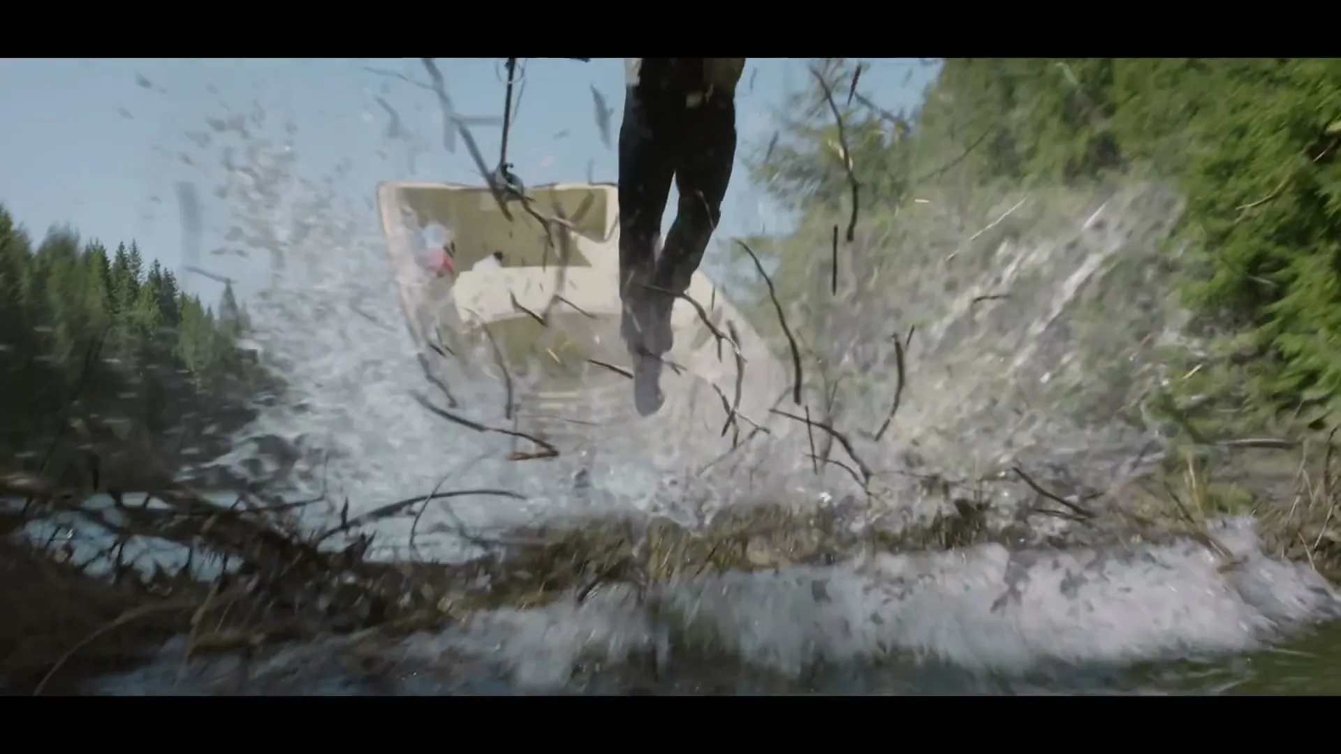 A man in a hat and vest jumps from a small boat, creating a large splash as he lands in a pile of branches in a lake surrounded by pine trees.