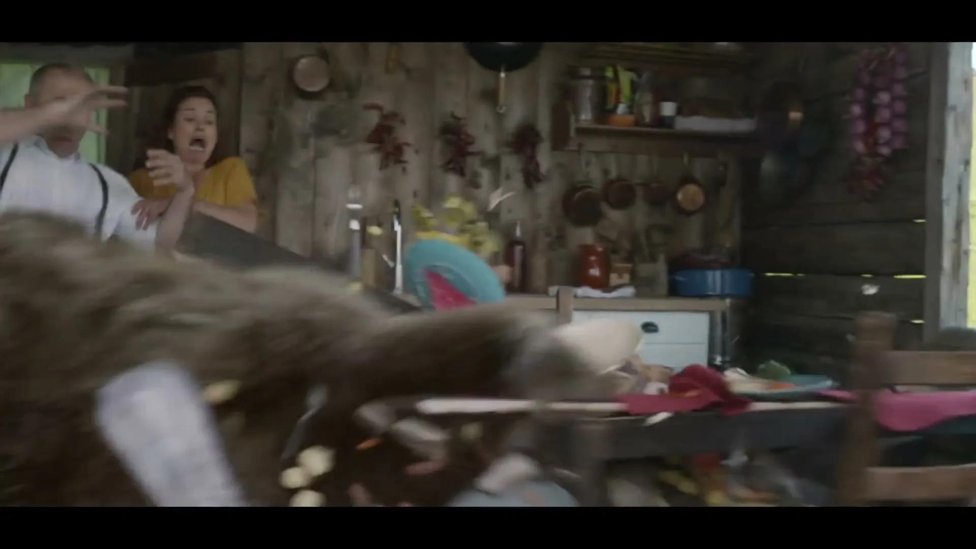 A man and woman are having dinner in a rustic cabin when a bear bursts through the door, causing chaos and sending the man flying out the window.