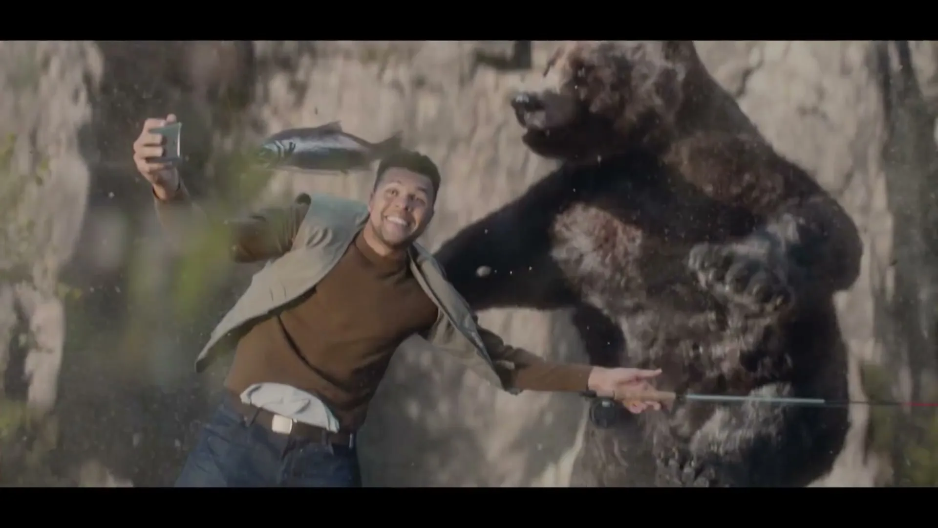 A man and a bear fall down a cliff, the man holding a phone and a fishing rod, with a fish on the line.