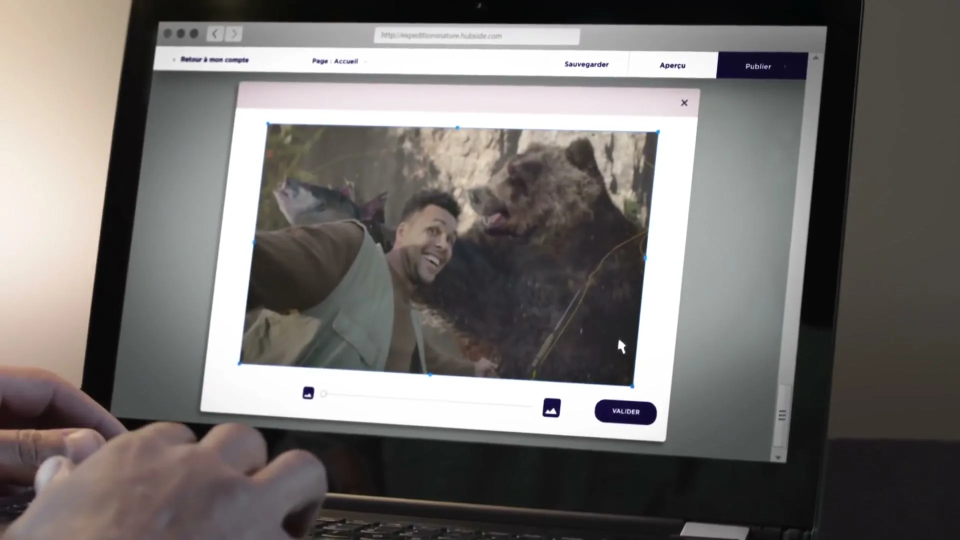 A person's hands are typing on a laptop, which displays a website editing interface with a photo of a man taking a selfie with a bear and a fish.