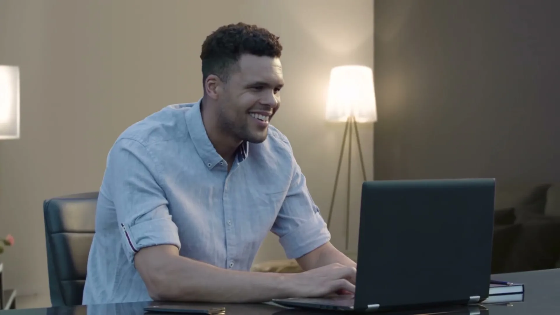 A man in his 30s sits at a desk, smiling and typing on a laptop in a modern interior.