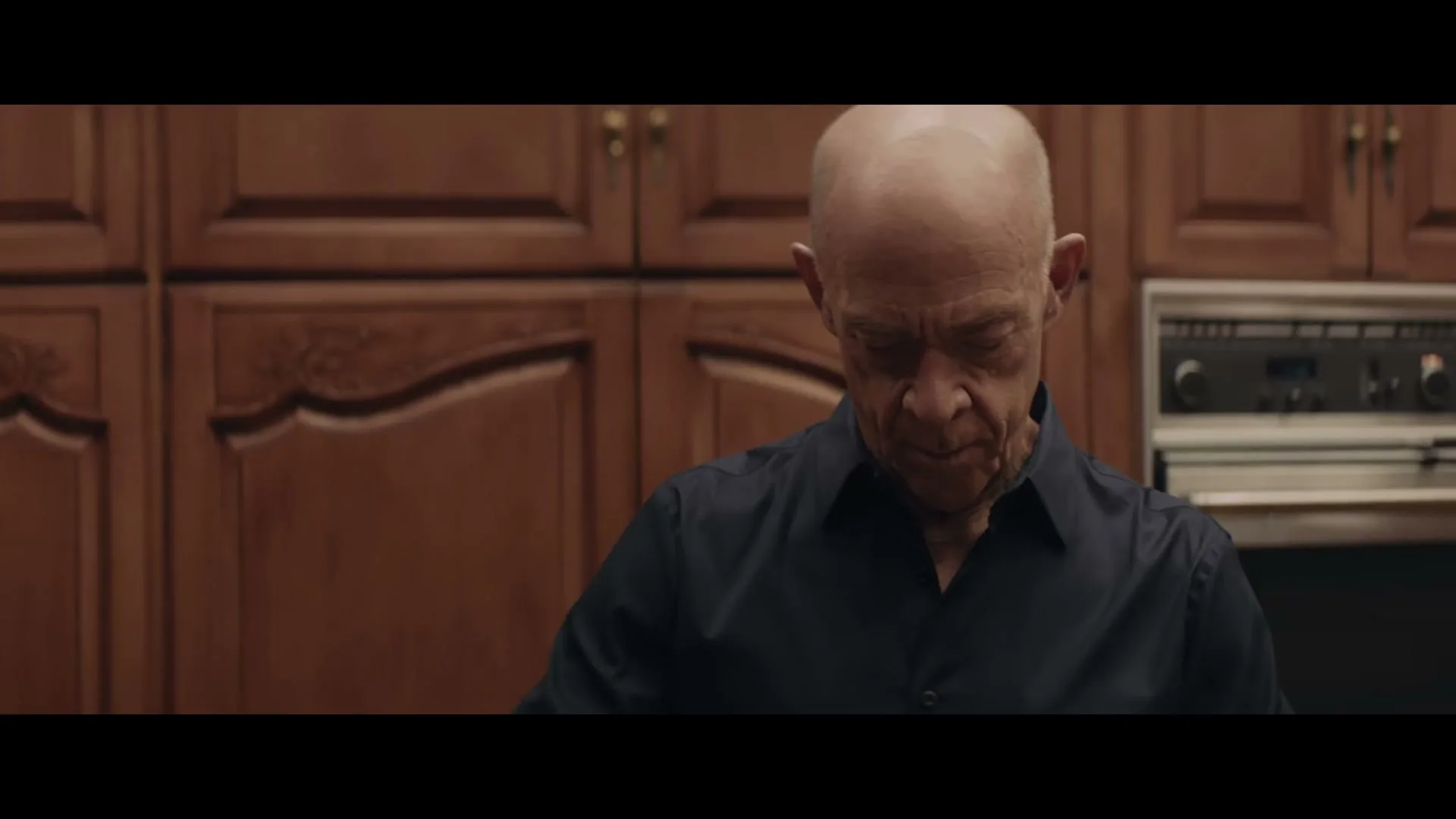 A bald man in his 60s, wearing a dark blue shirt, looks down with a serious expression in a kitchen with wooden cabinets.
