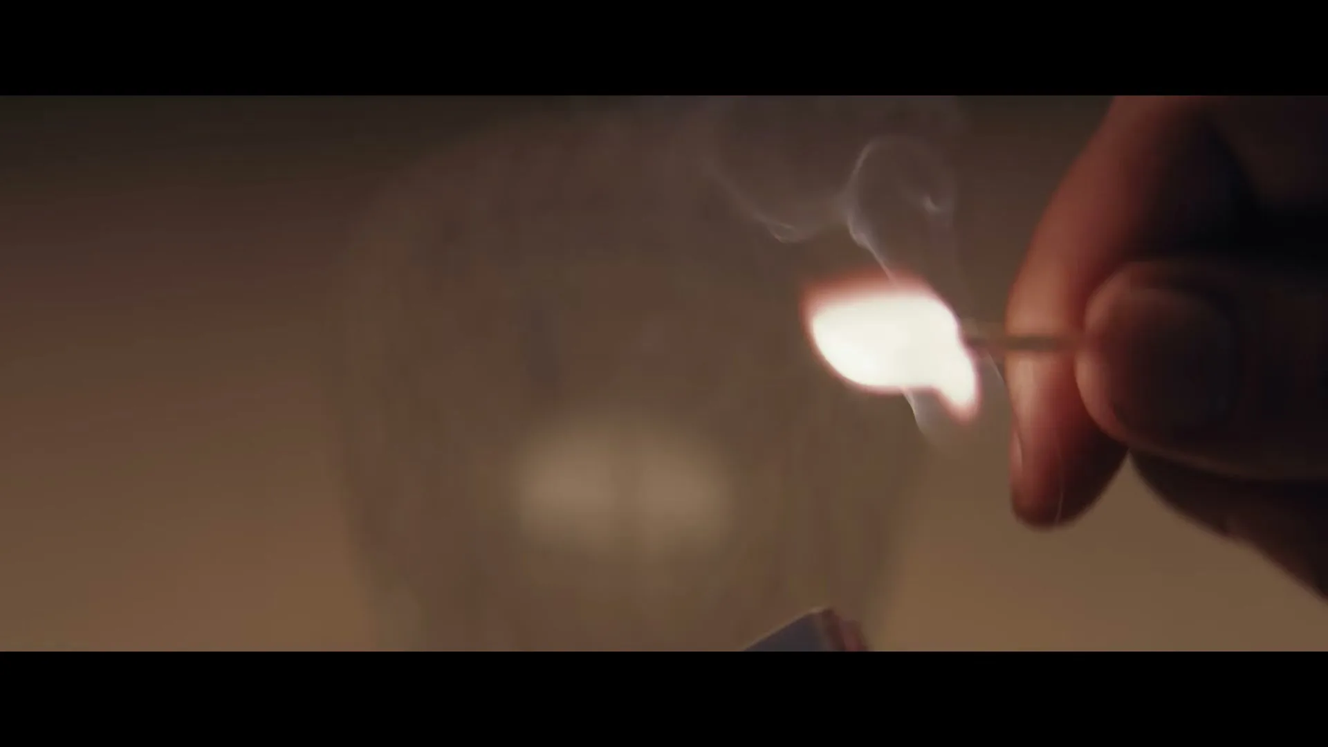 A person's hand strikes a match, creating a small flame and smoke, with a blurred background.