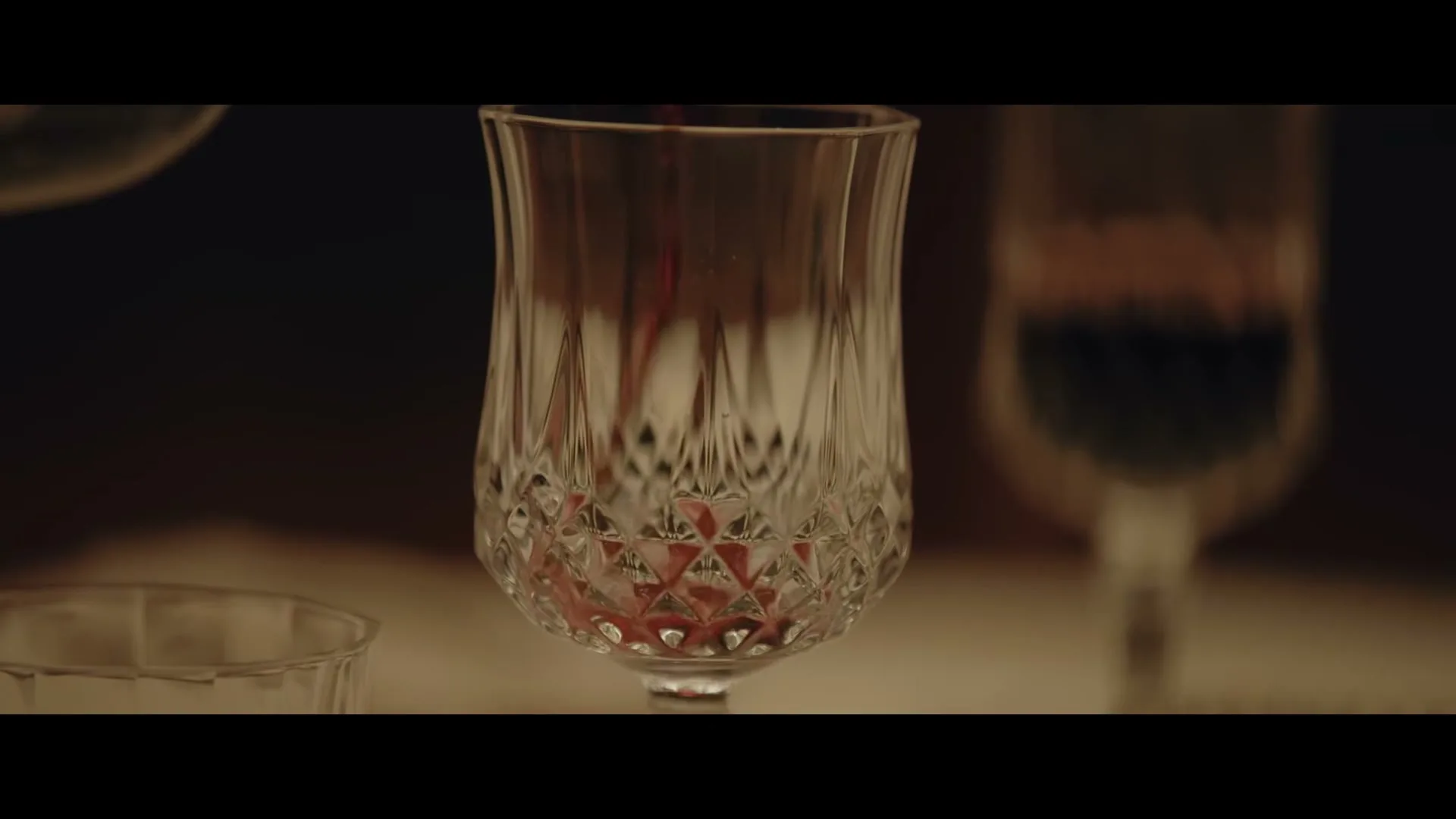 Red liquid is poured into a crystal glass on a table, with other glasses blurred in the background, creating an intimate atmosphere.