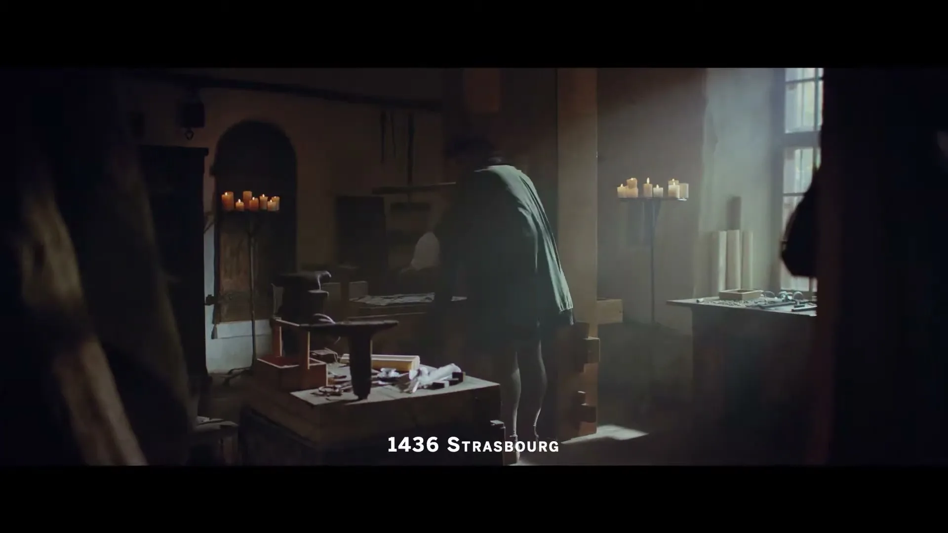A man in medieval clothing works at a wooden table in a dimly lit workshop, illuminated by sunlight and candles.
