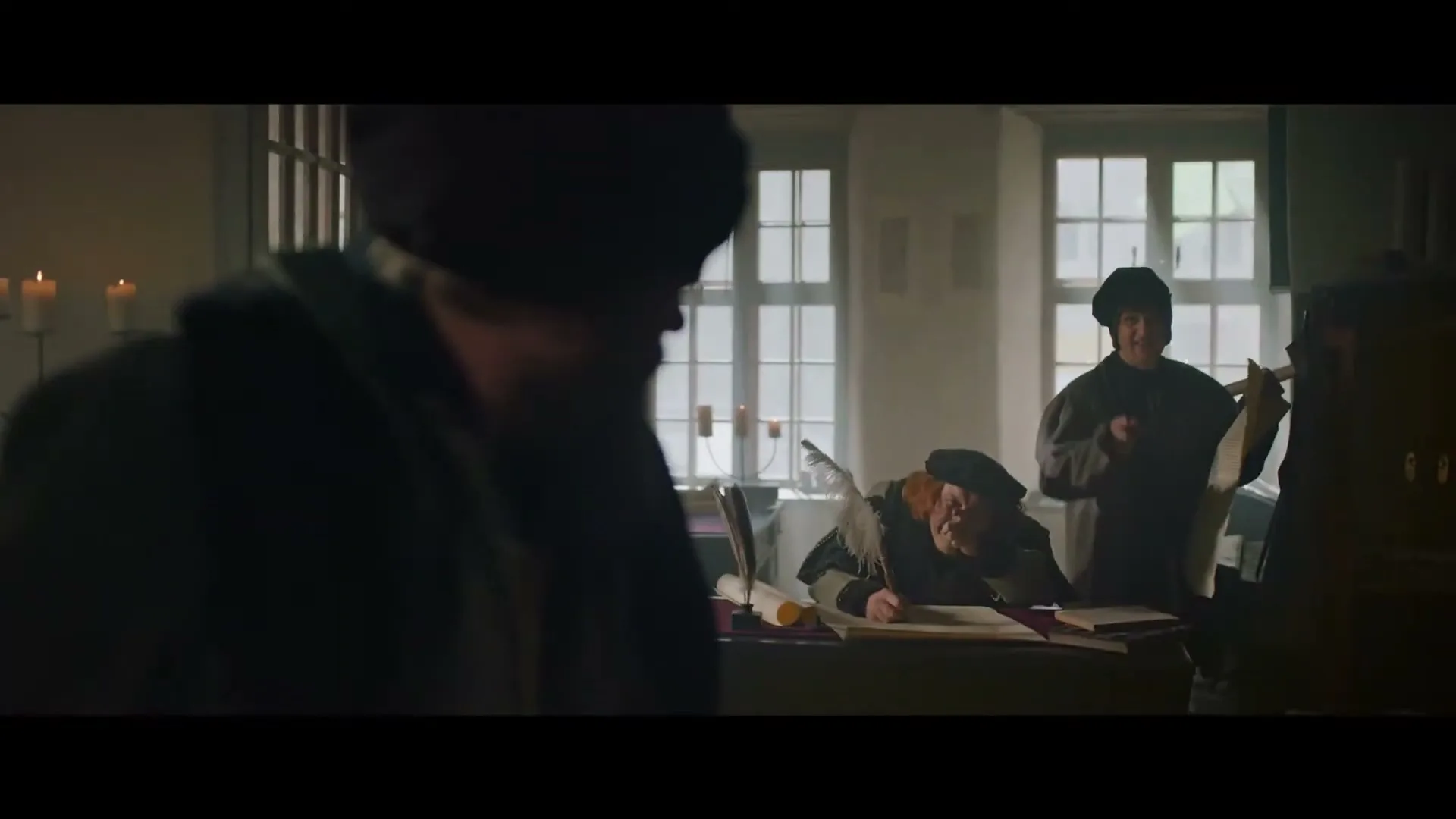 A man with a long beard and a dark hat looks at a large, old document in a dimly lit room with other men in the background.