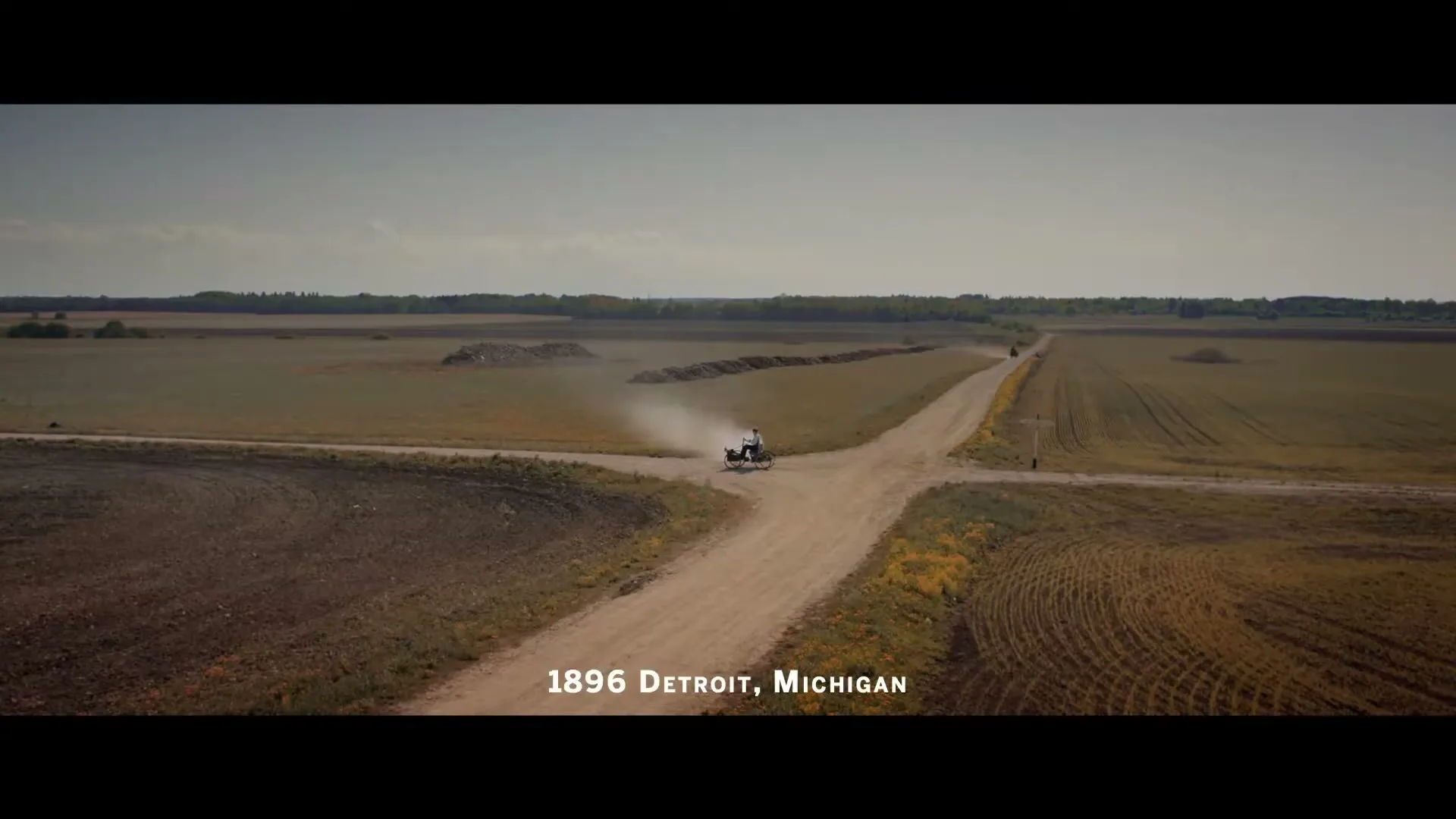 An early automobile, a quadricycle, drives along a dusty dirt road through vast, flat fields under a bright sky, with text "1896 Detroit, Michigan" on screen.