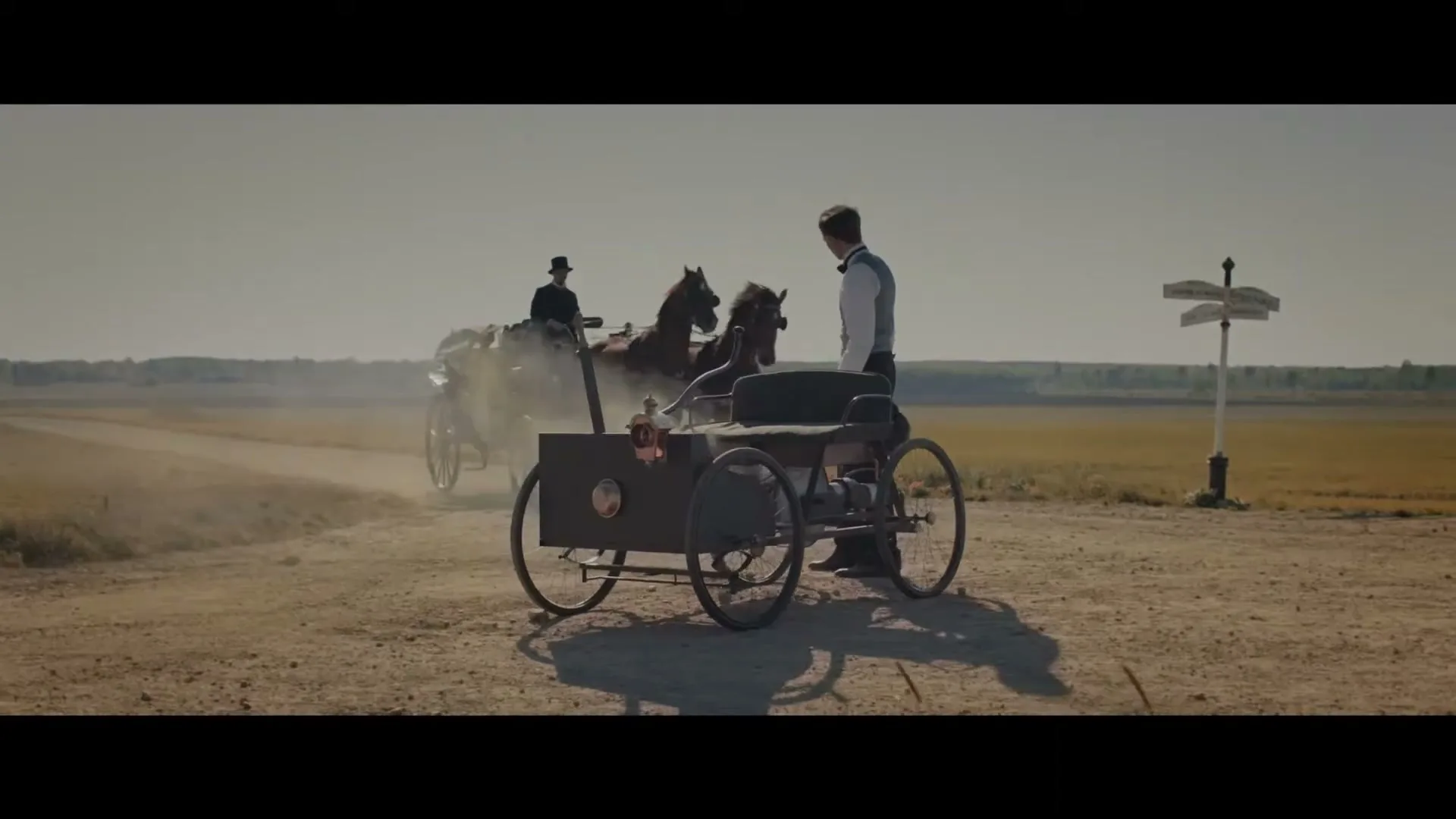 A man in a white shirt and vest stands next to an early automobile on a dirt road, as a horse-drawn carriage passes by, kicking up dust in a wide-open field.
