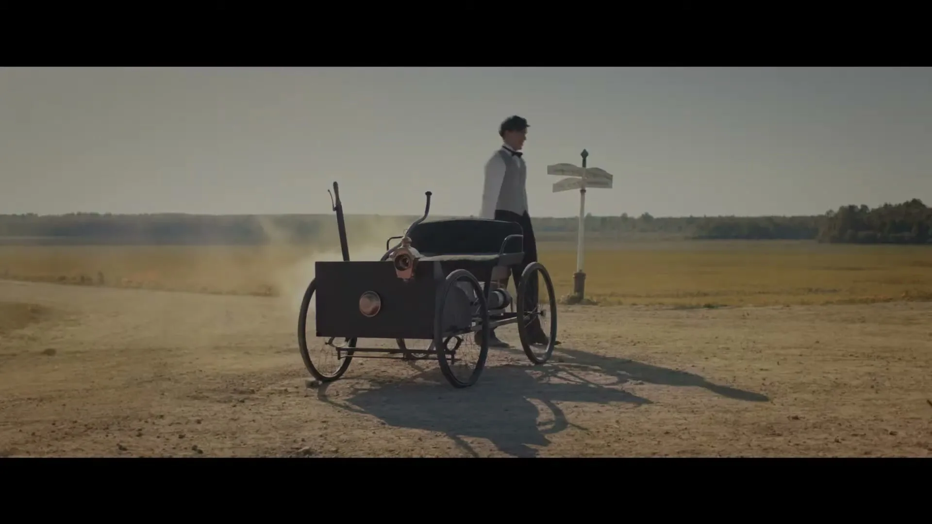 A young man in vintage attire attempts to start an old-fashioned car on a dusty rural road, with a signpost nearby.