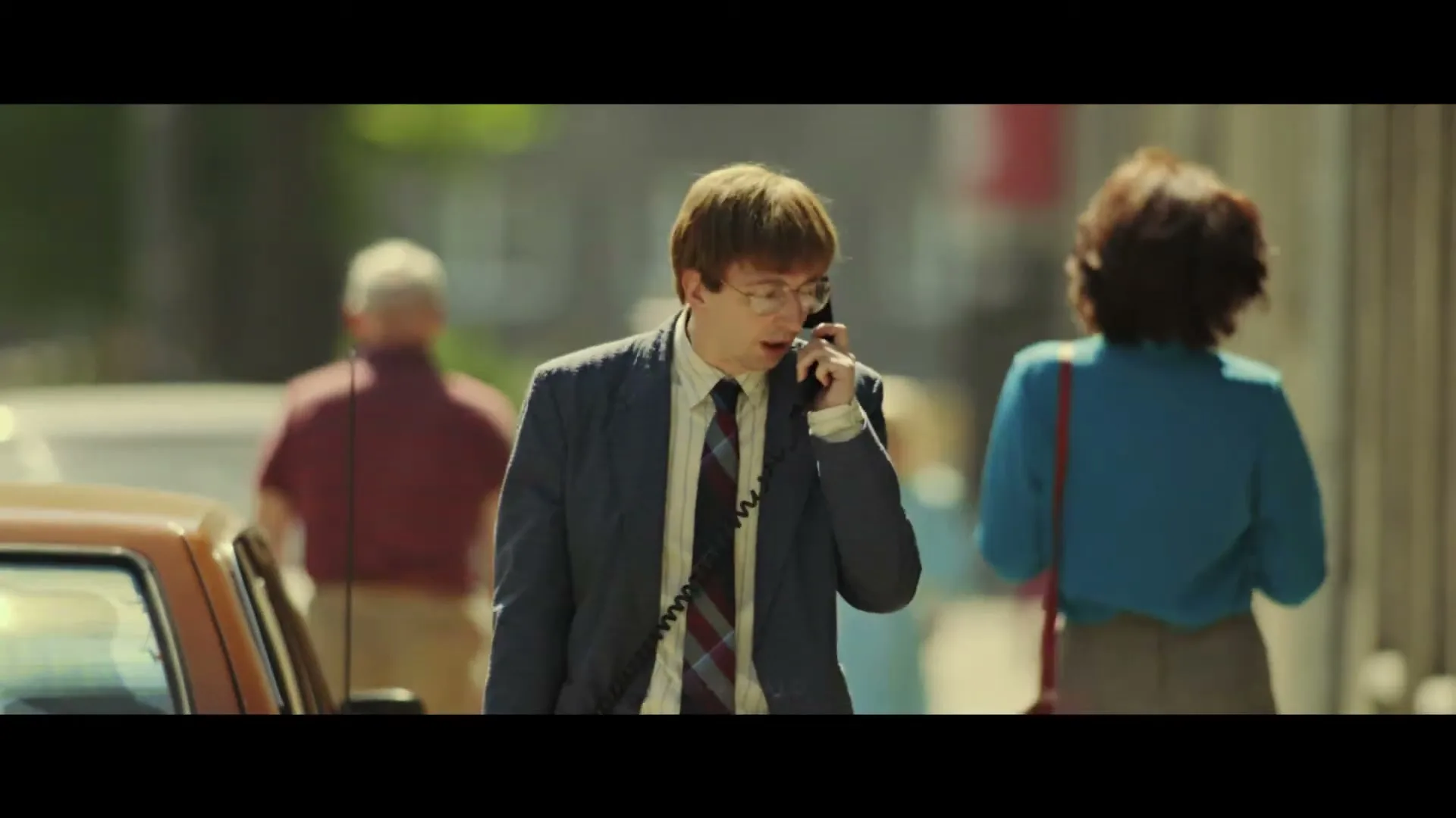 A young man in a suit and glasses walks down a sunny street, holding a large, old-fashioned phone to his ear, looking confused.