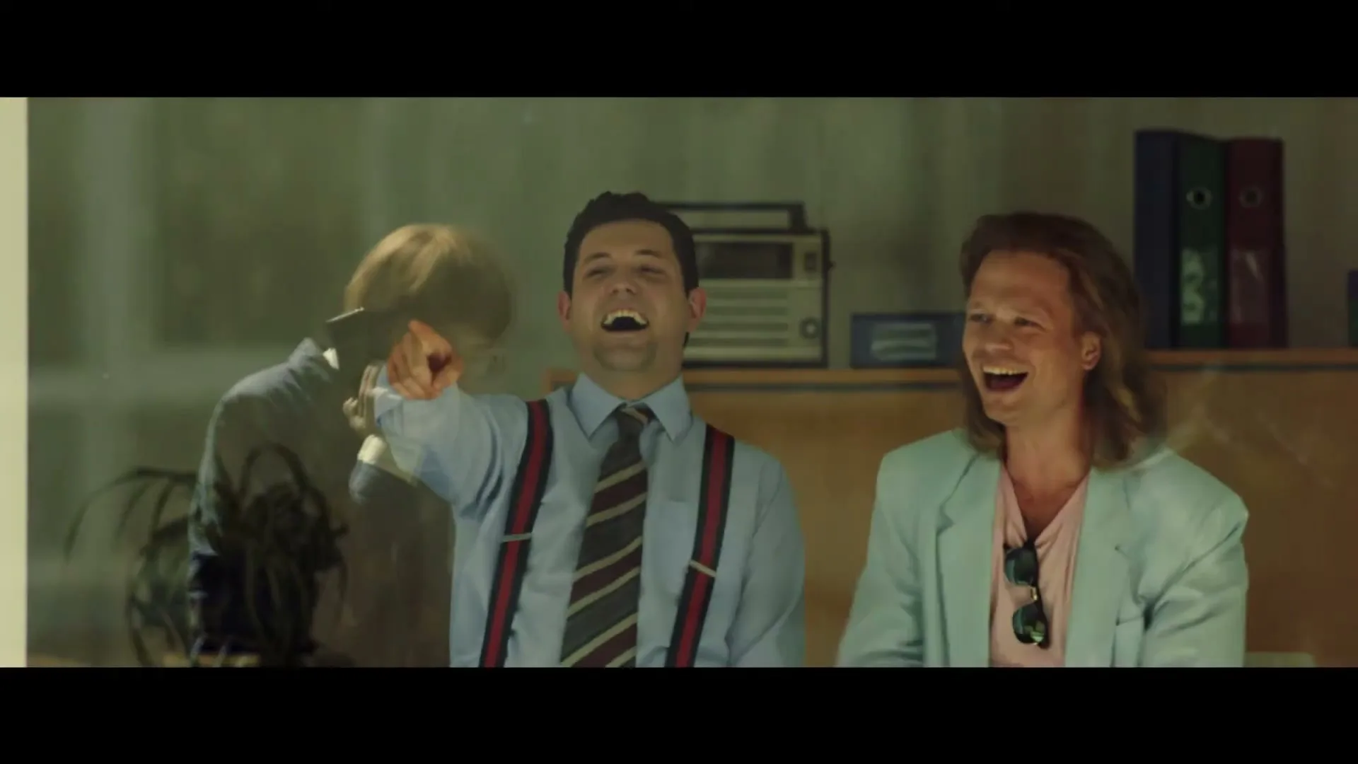 Two men, one in a shirt and suspenders, the other in a light blue jacket, laugh hysterically in an office, one holding a phone.