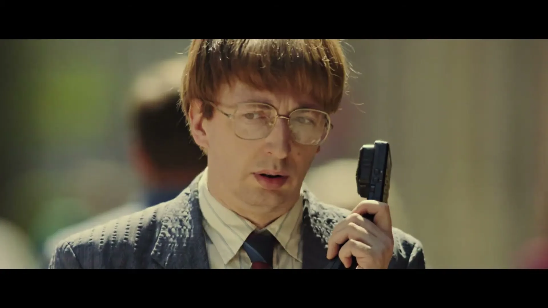 A man with a bowl cut and glasses holds a walkie-talkie, looking around with a concerned expression in a sunny outdoor setting.