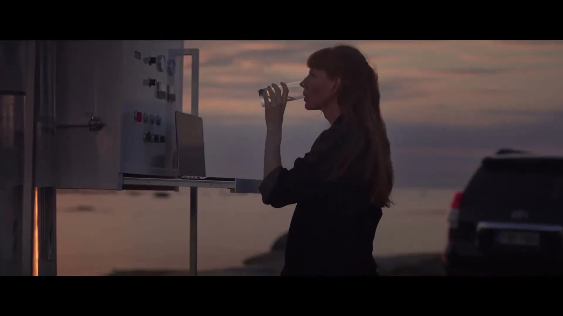 A woman with long red hair in a dark shirt drinks from a glass next to a metal box with a laptop, with a car and the sea in the background at dusk.
