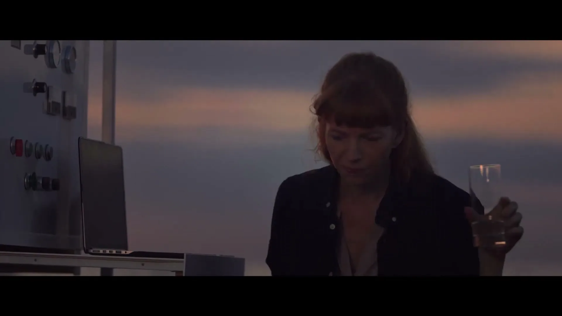 A woman with long red hair drinks water while looking at a laptop on a rooftop at dusk, with text appearing on screen.