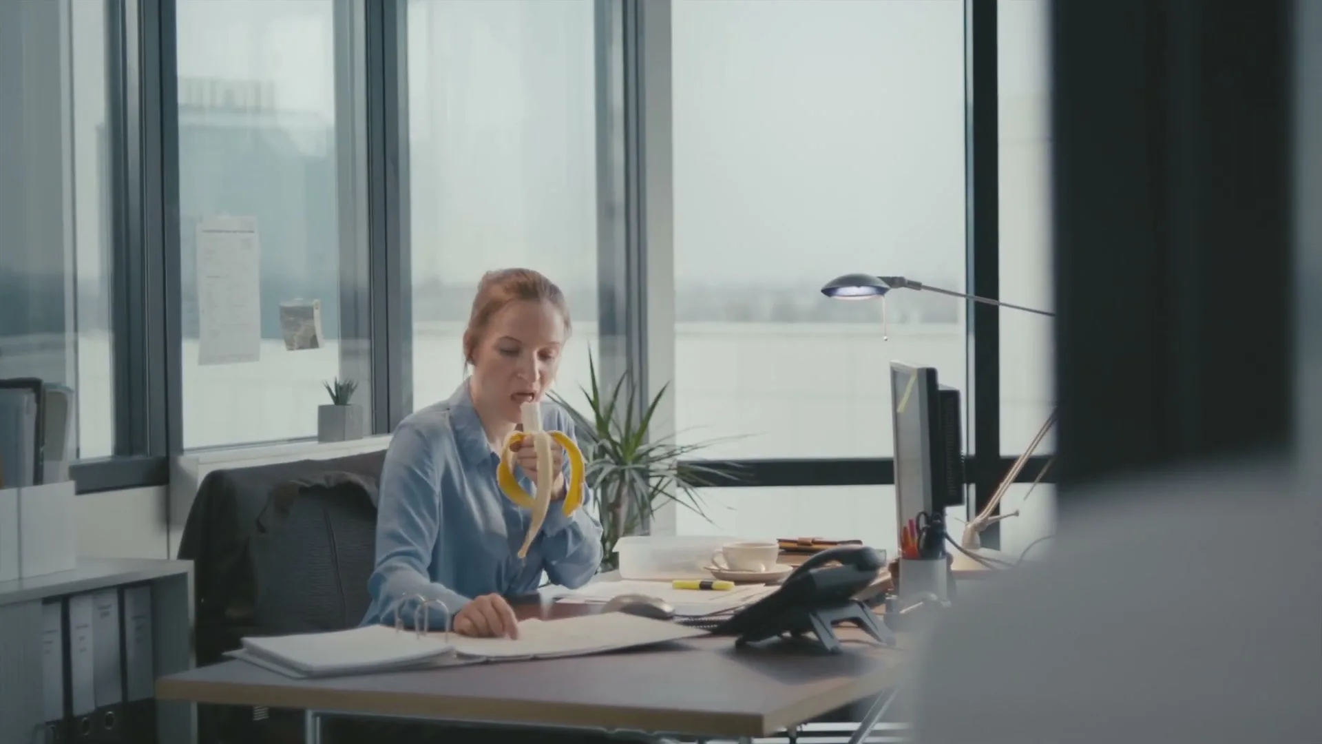 A young woman with her hair in a bun, wearing a light blue shirt, eats a banana at her desk in a modern office with large windows.